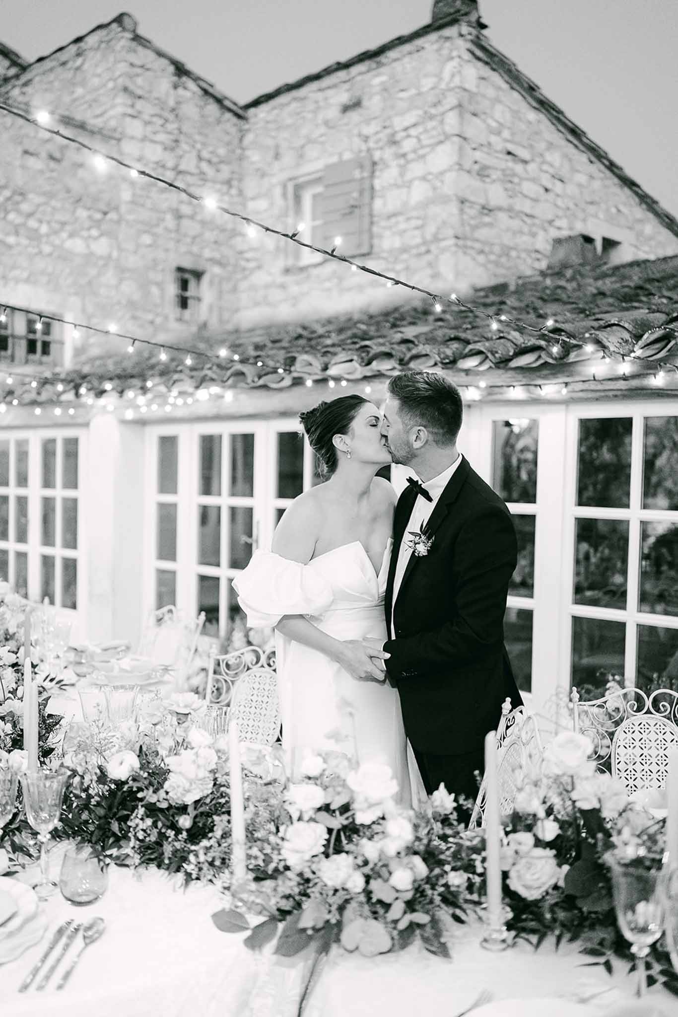 Bride and groom kissing during reception dinner in courtyard with stone cottage and pavilion backdrop