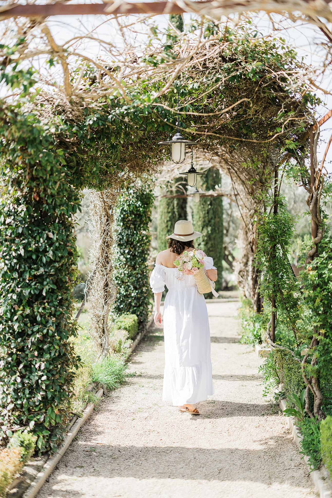 Bride walking down garden pathway in white dress and straw hat carrying blush bouquet at outdoor wedding venue