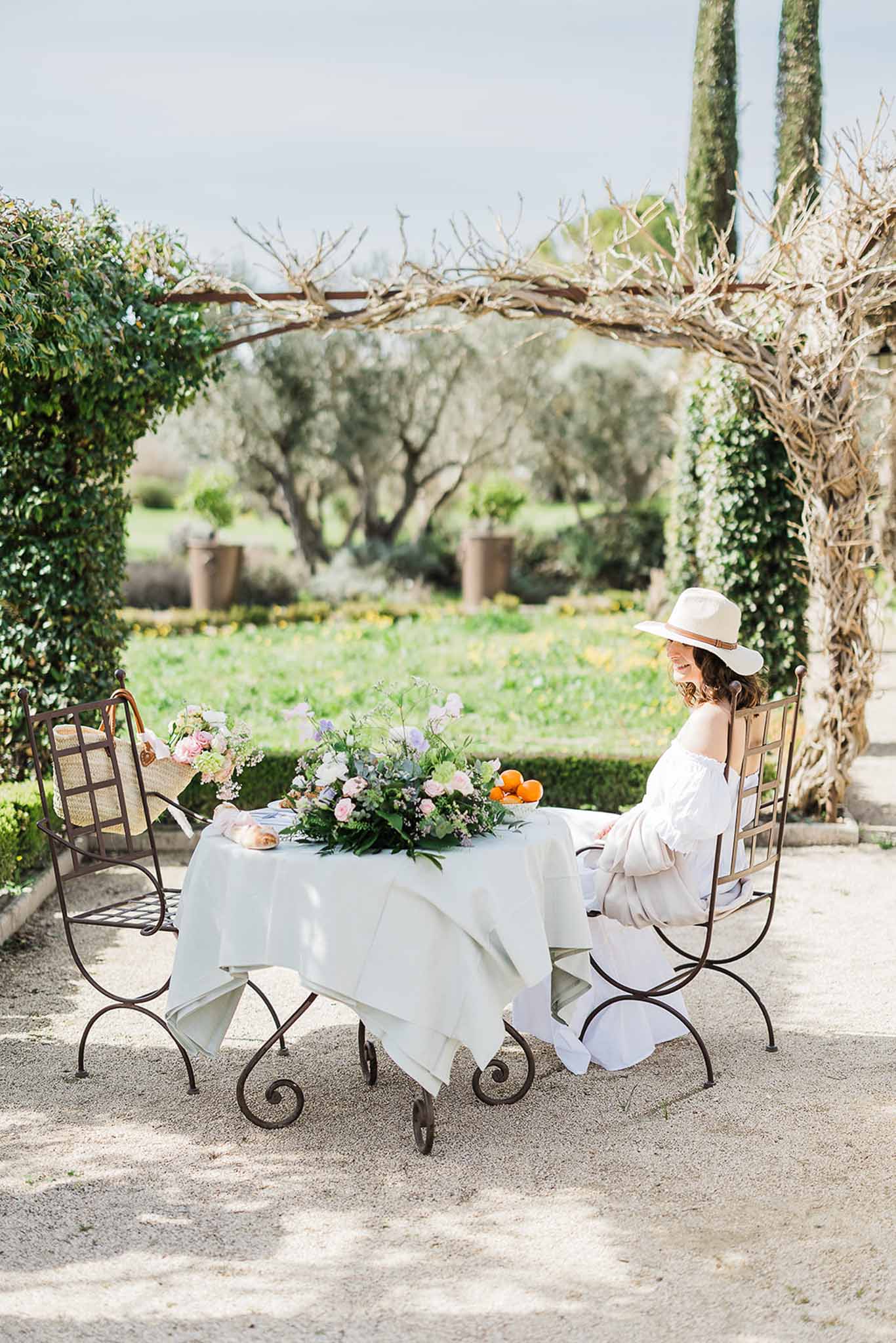 Bride in white dress and straw hat seated at garden table with floral arrangement in Mediterranean setting