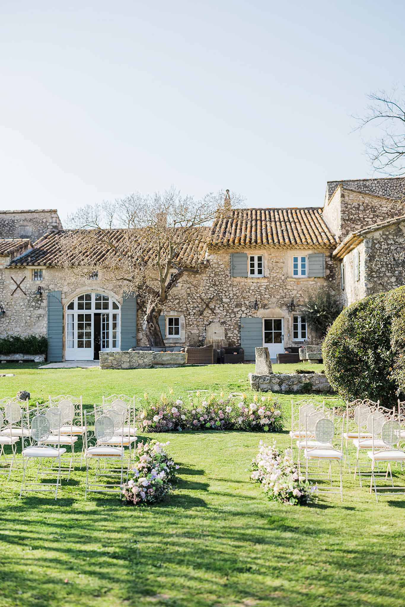 Outdoor wedding ceremony setup on lawn in front of historic French stone farmhouse with white chairs and floral arrangements