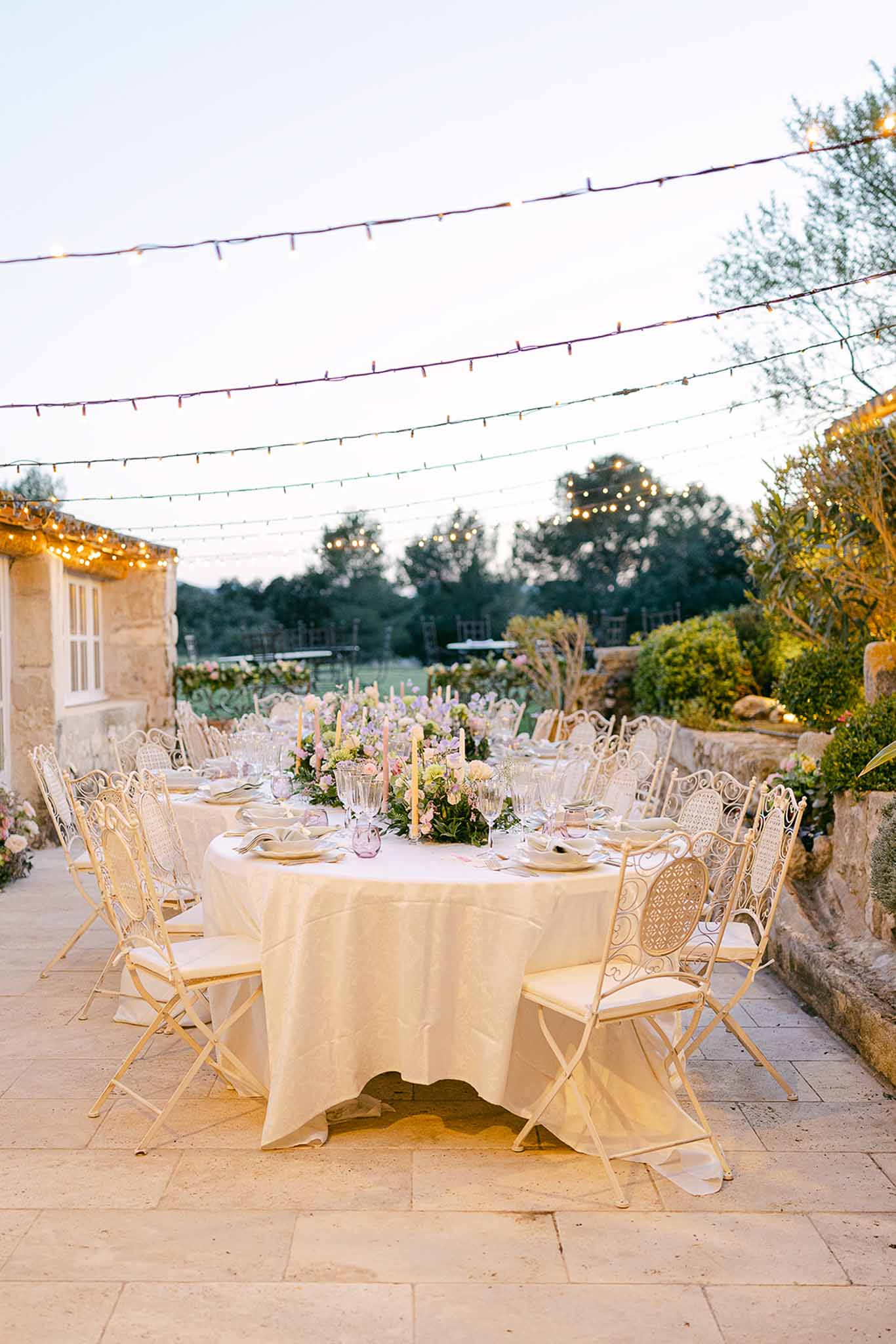 Outdoor reception dinner setup with round tables and string lights on stone terrace at countryside venue