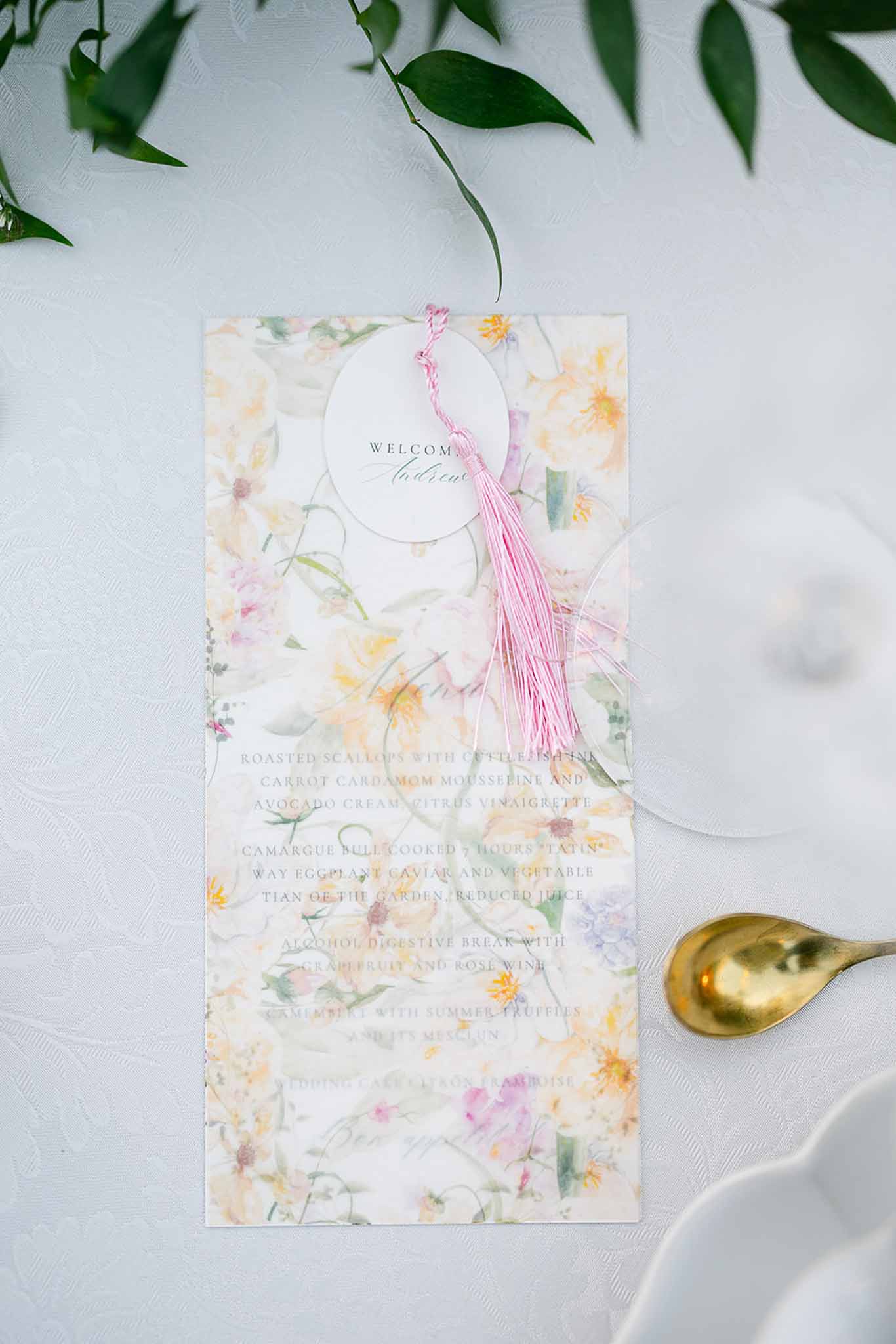 Wedding place setting with watercolor floral menu card, pink tassel name tag, and gold spoon on white linen