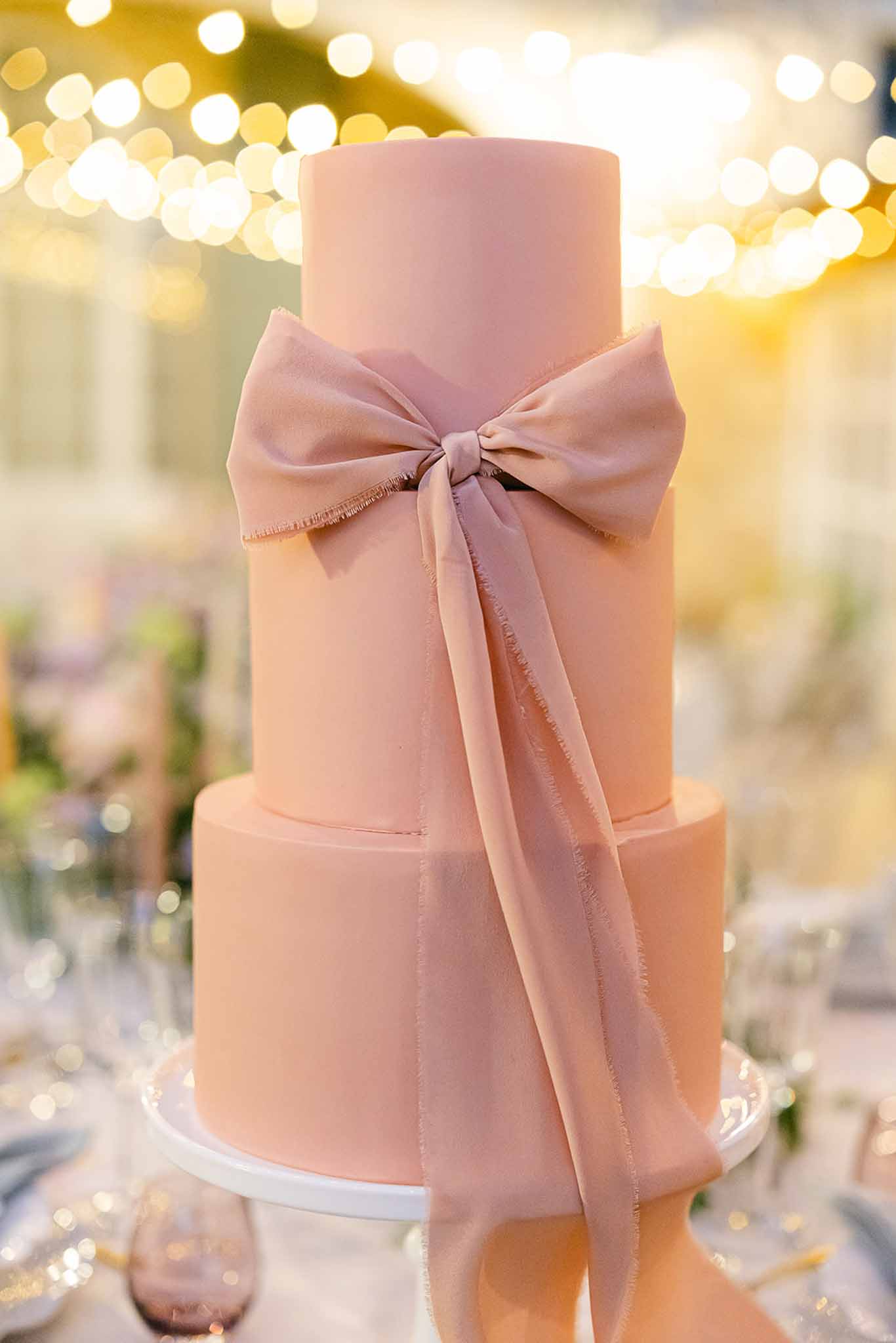 Two-tiered wedding cake with dusty rose frosting and ribbon bow at indoor reception with string lights