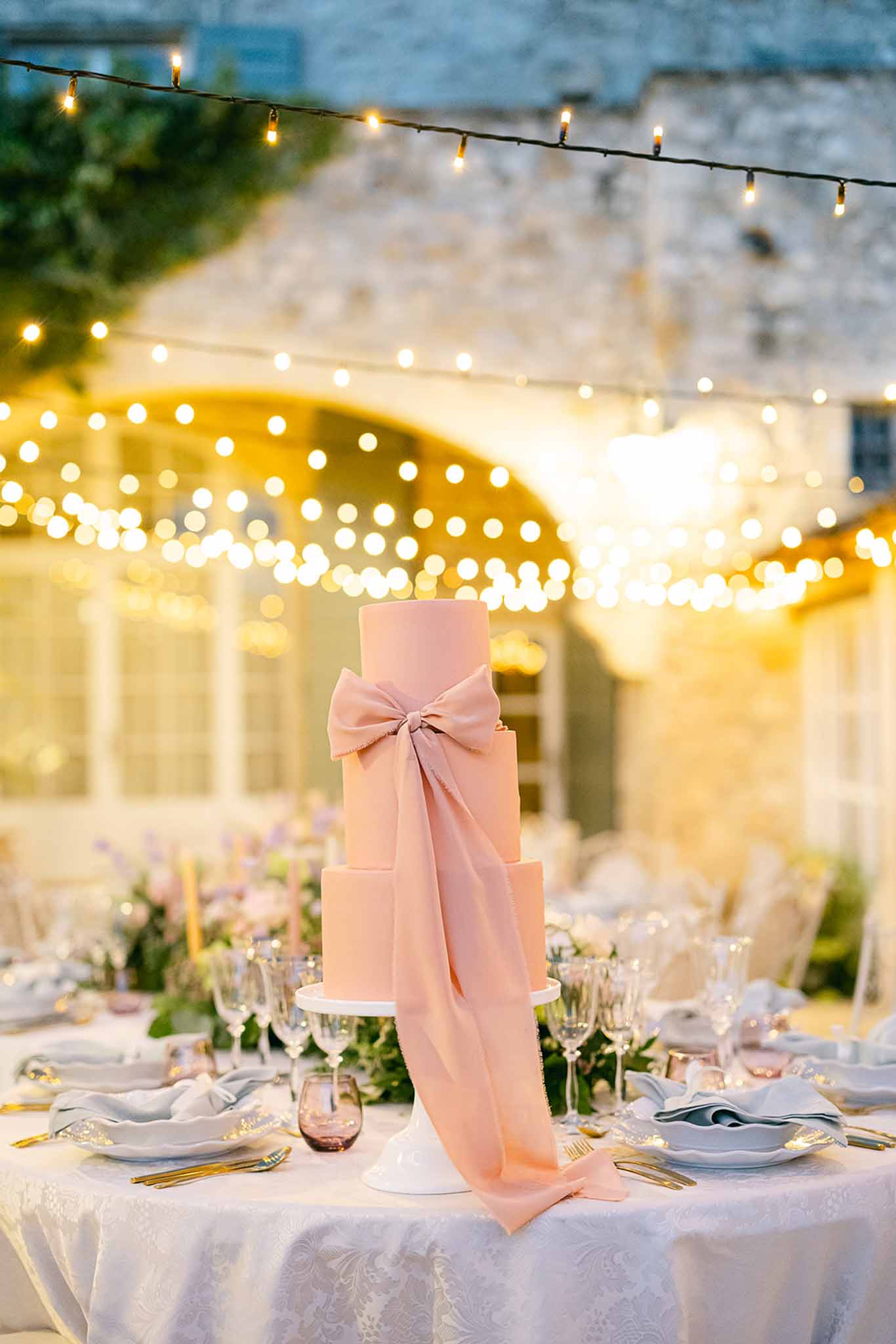 Three-tiered wedding cake with dusty rose fondant and gold table setting at outdoor reception venue