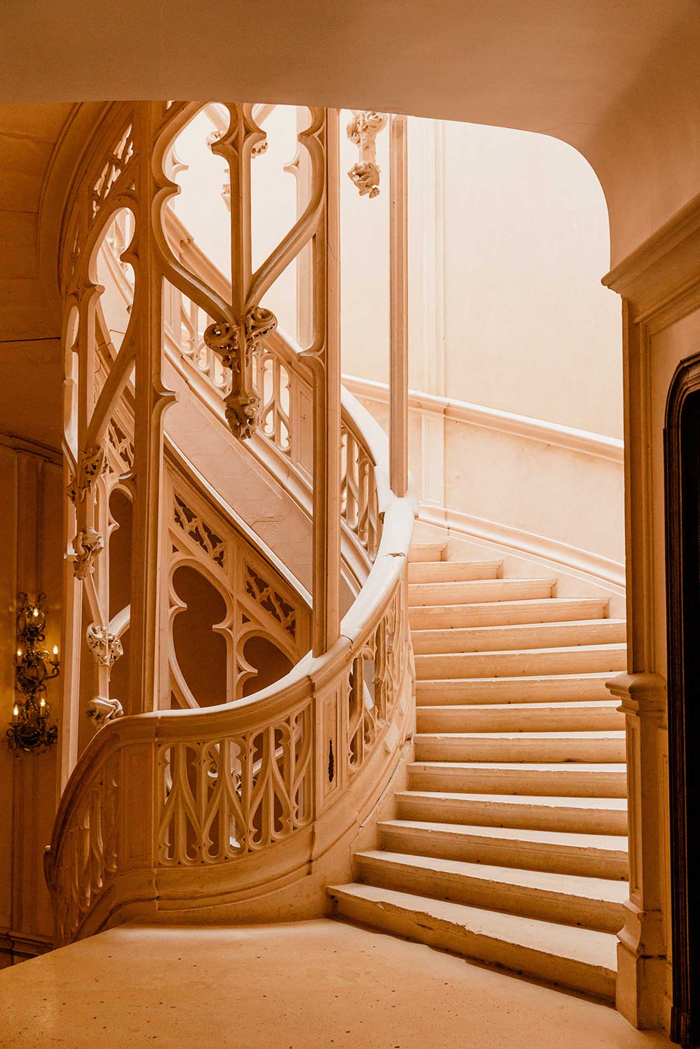 Grand sweeping Gothic-style carved stone staircase with trefoil balustrades inside a French chateau lit by warm sconces