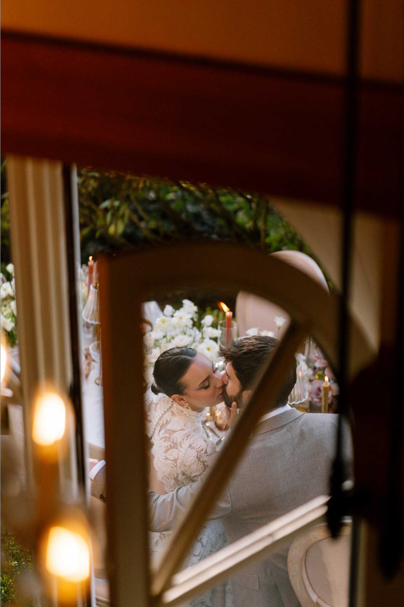Bride and groom kissing at altar through decorative wooden frame with candles and white floral arrangements