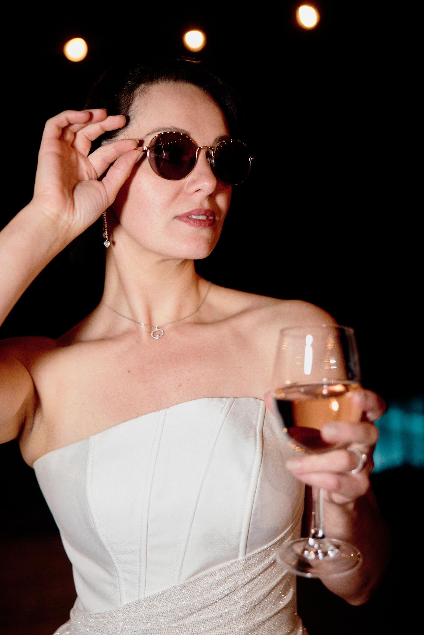 Bride in strapless corseted gown holding rose wine and adjusting gold sunglasses under spotlights at evening reception