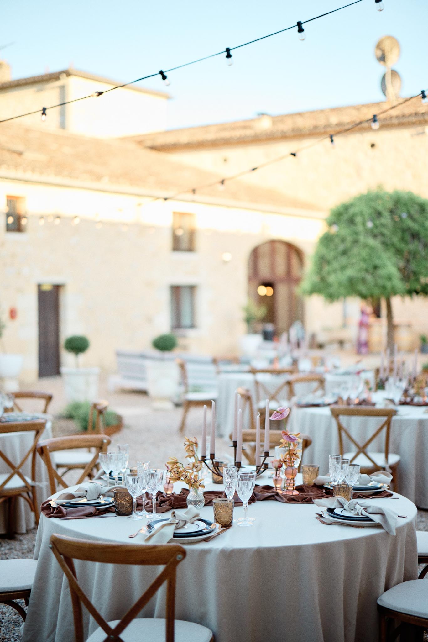 Round tables with terracotta runners, black candelabras, and dusty pink candles under string lights in courtyard