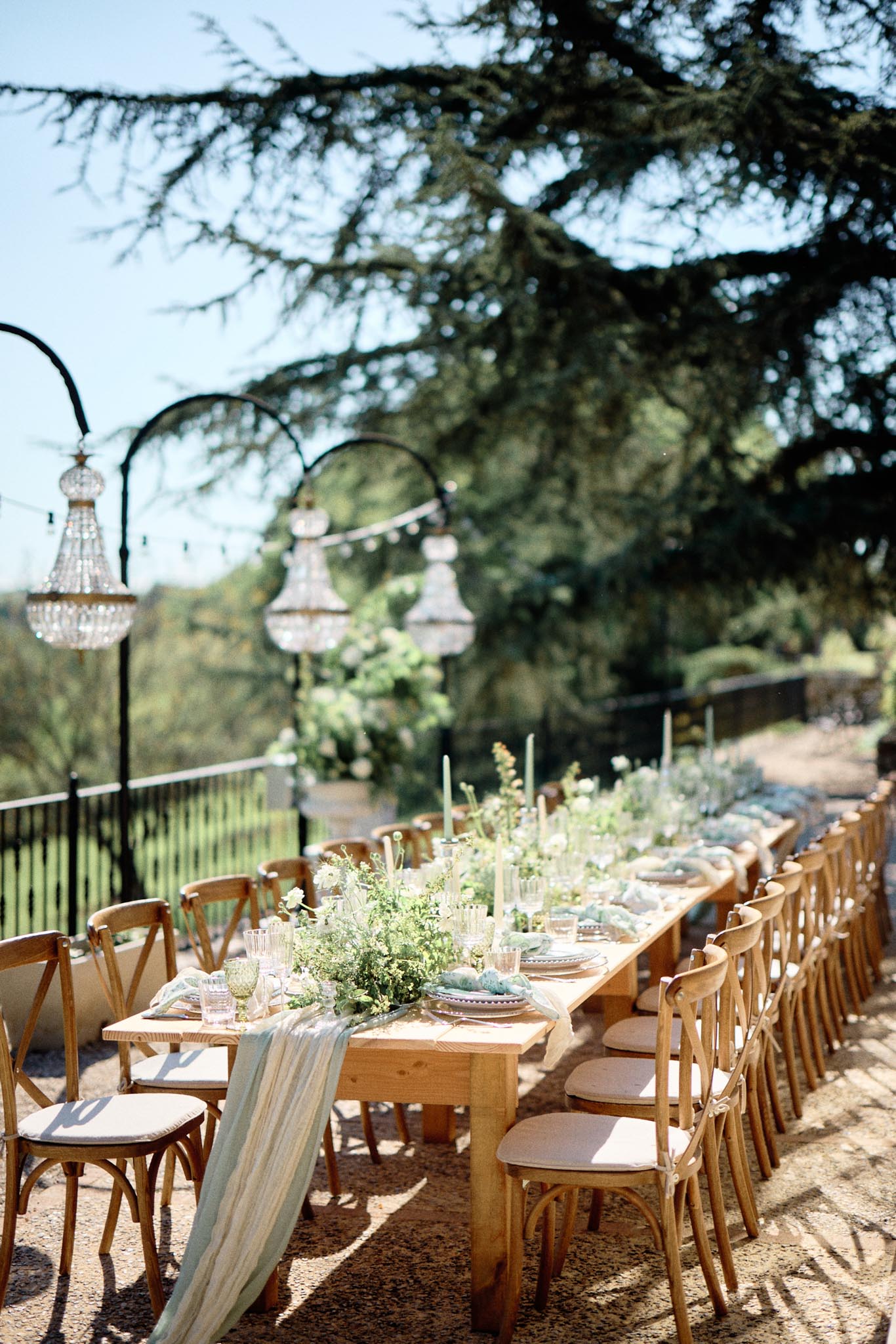 Long farm table reception setup with sage green runner, greenery centerpiece, chandeliers, and cross-back chairs on gravel...