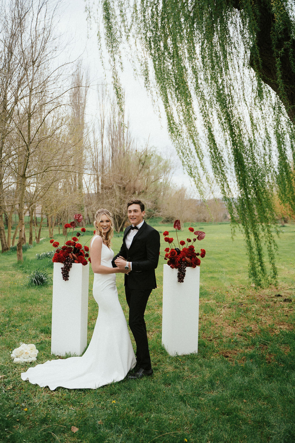 Bride in ivory satin gown and groom in black tuxedo between white plinths with deep red floral arrangements