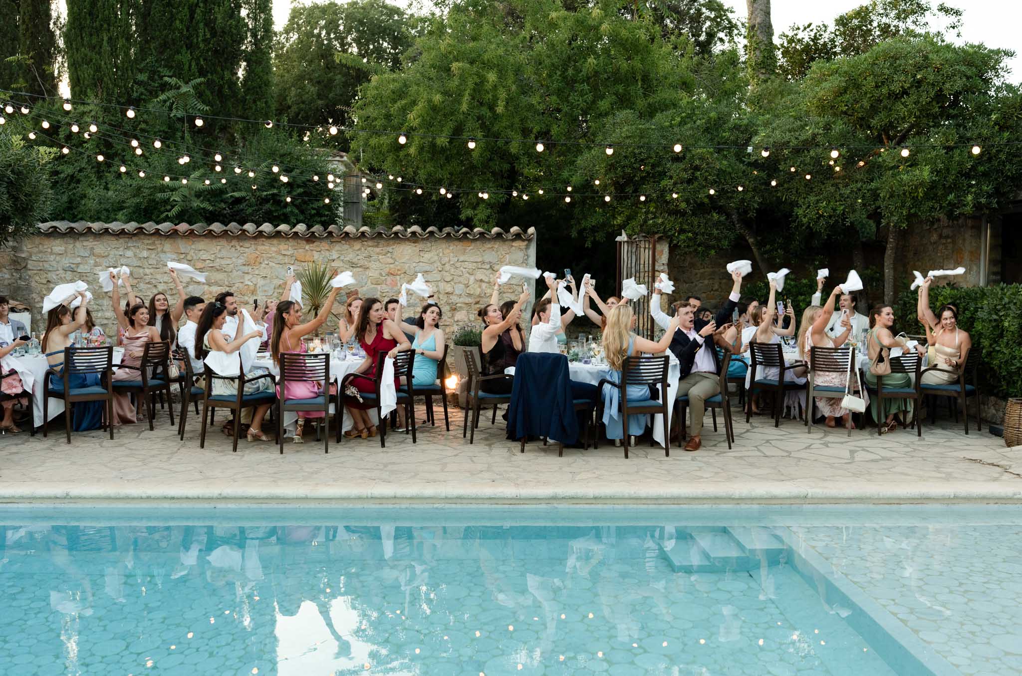 Poolside reception dinner with guests waving white napkins under fairy lights at Provencal terrace venue