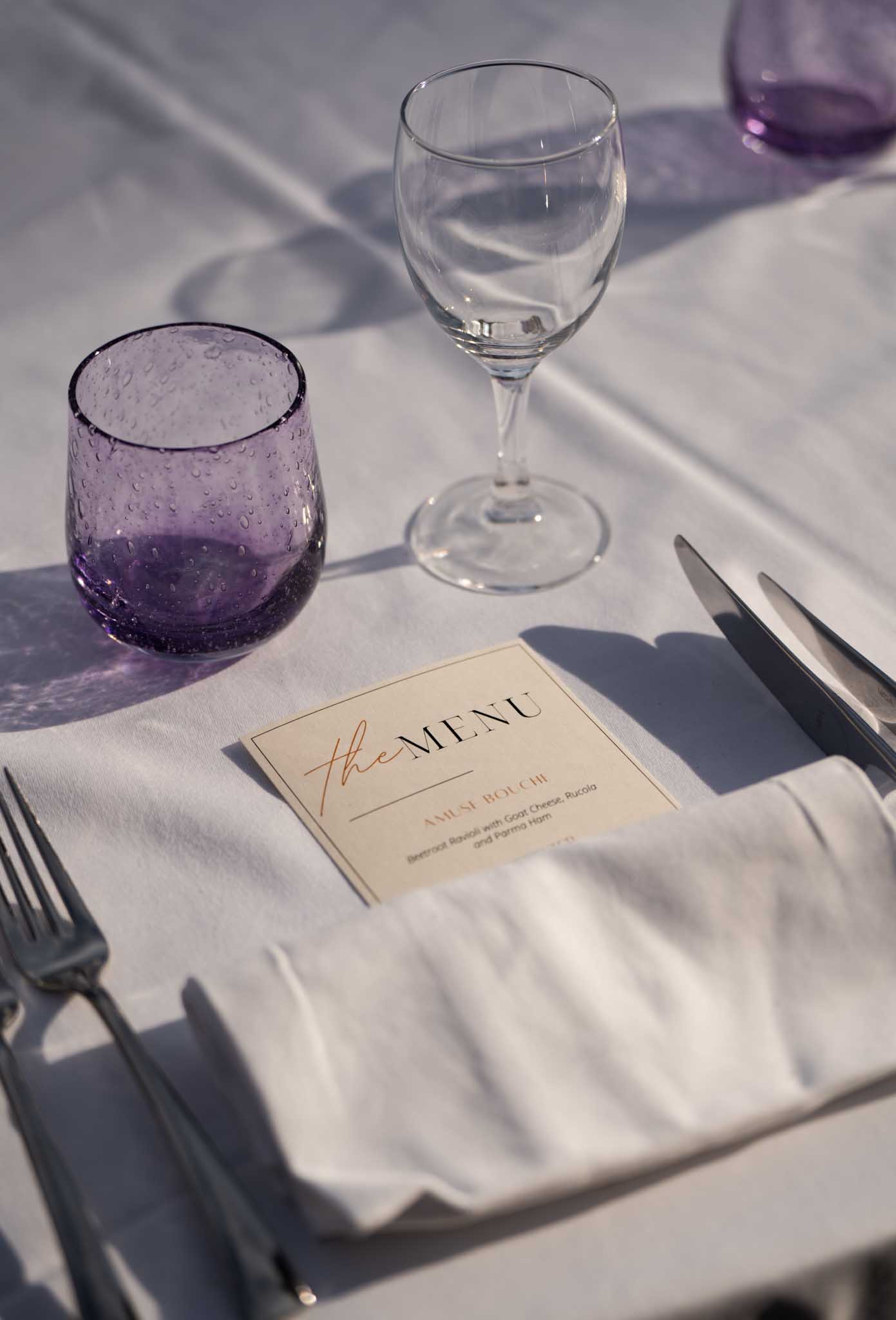 White linen place setting with printed menu card and amethyst purple bubble glass tumblers in outdoor reception