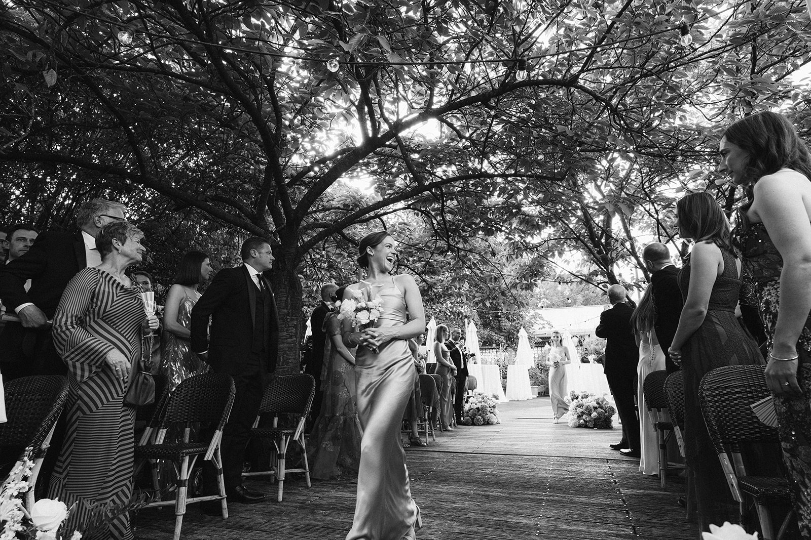 Black and white wide view of the bride walking the aisle beneath trees at Restaurant l'Ile, Paris