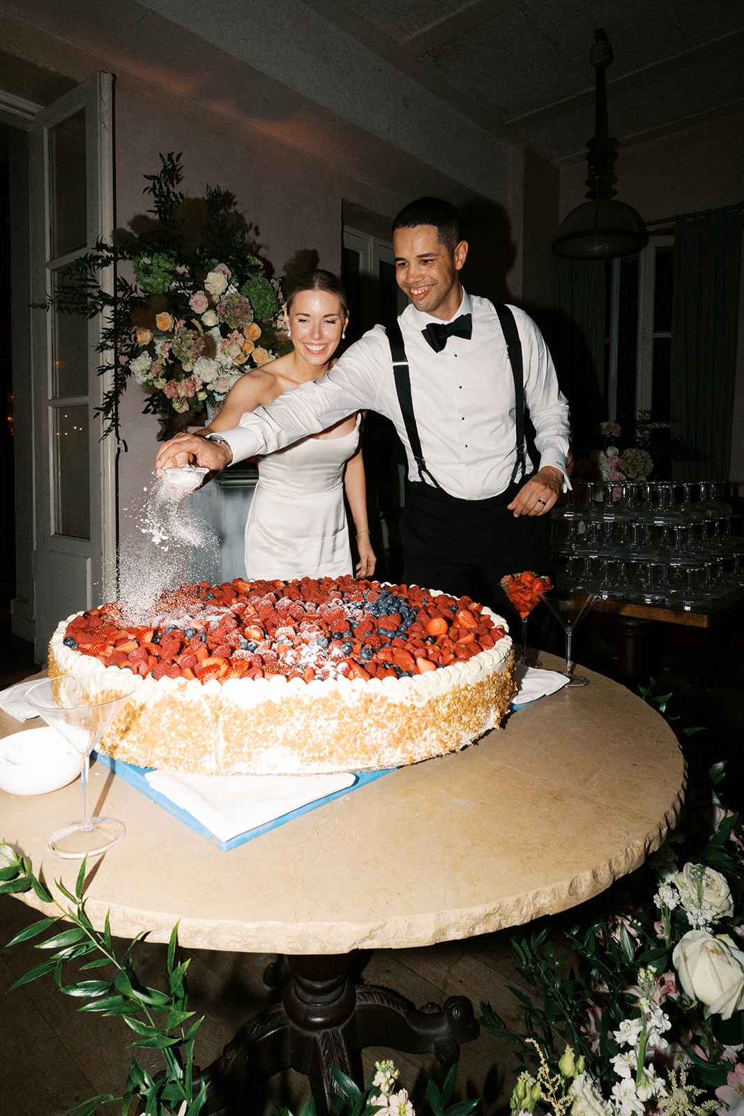 Couple cutting a strawberry-topped cake on a stone table with a champagne tower behind