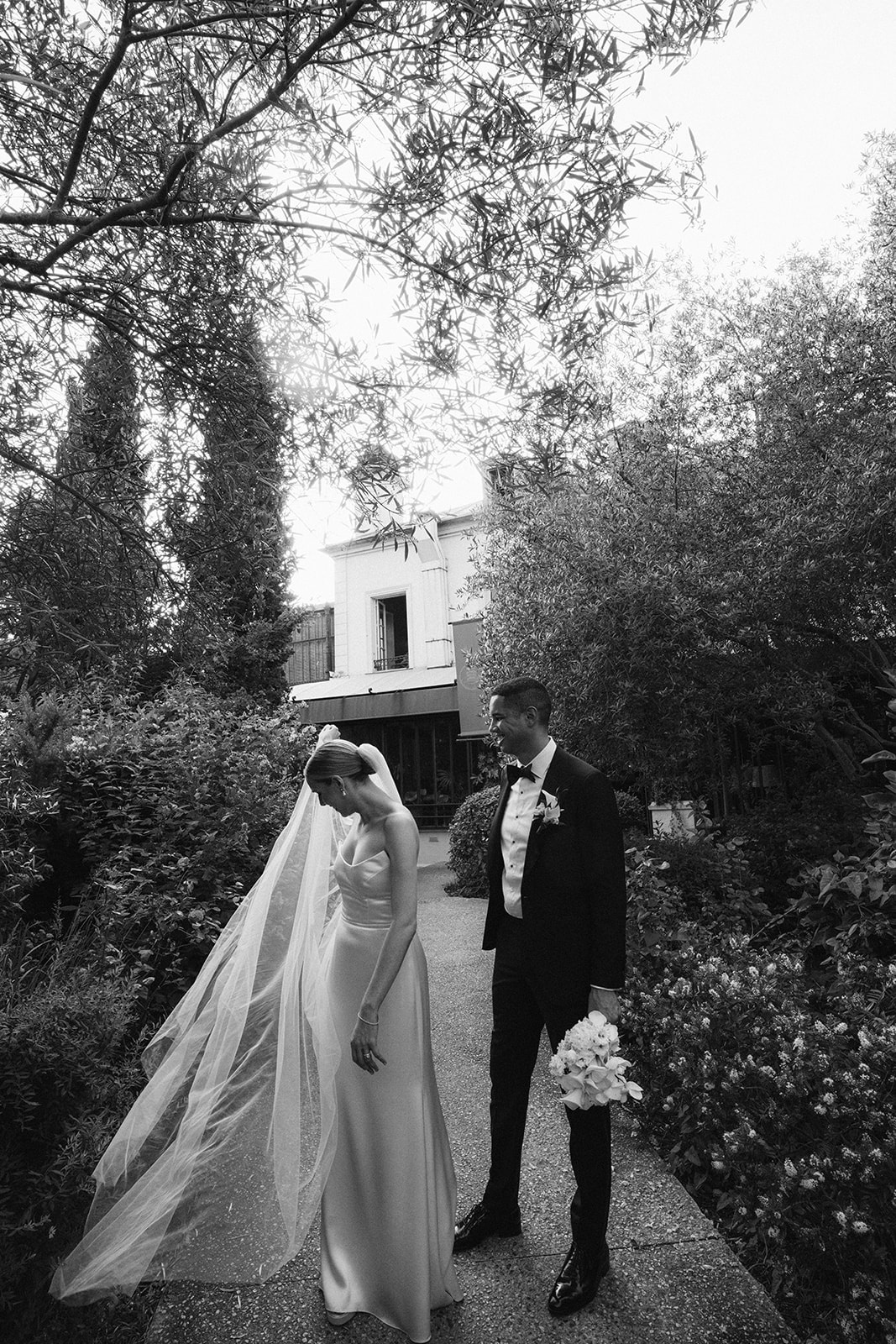 Black and white portrait of the couple in the venue garden with the bride's veil flowing behind