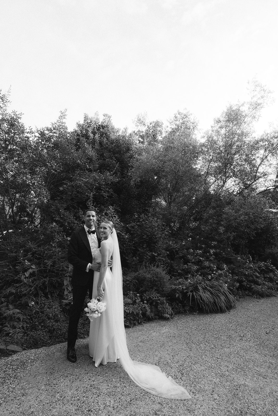 Black and white portrait of the couple smiling in a leafy garden, the bride's veil pooling behind