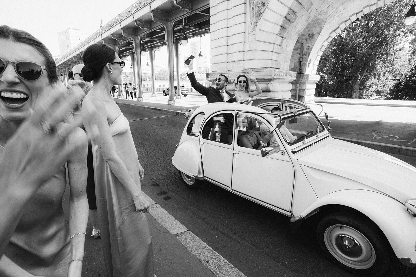 Black and white shot of guests waving at a vintage Citroen 2CV beneath a Paris bridge