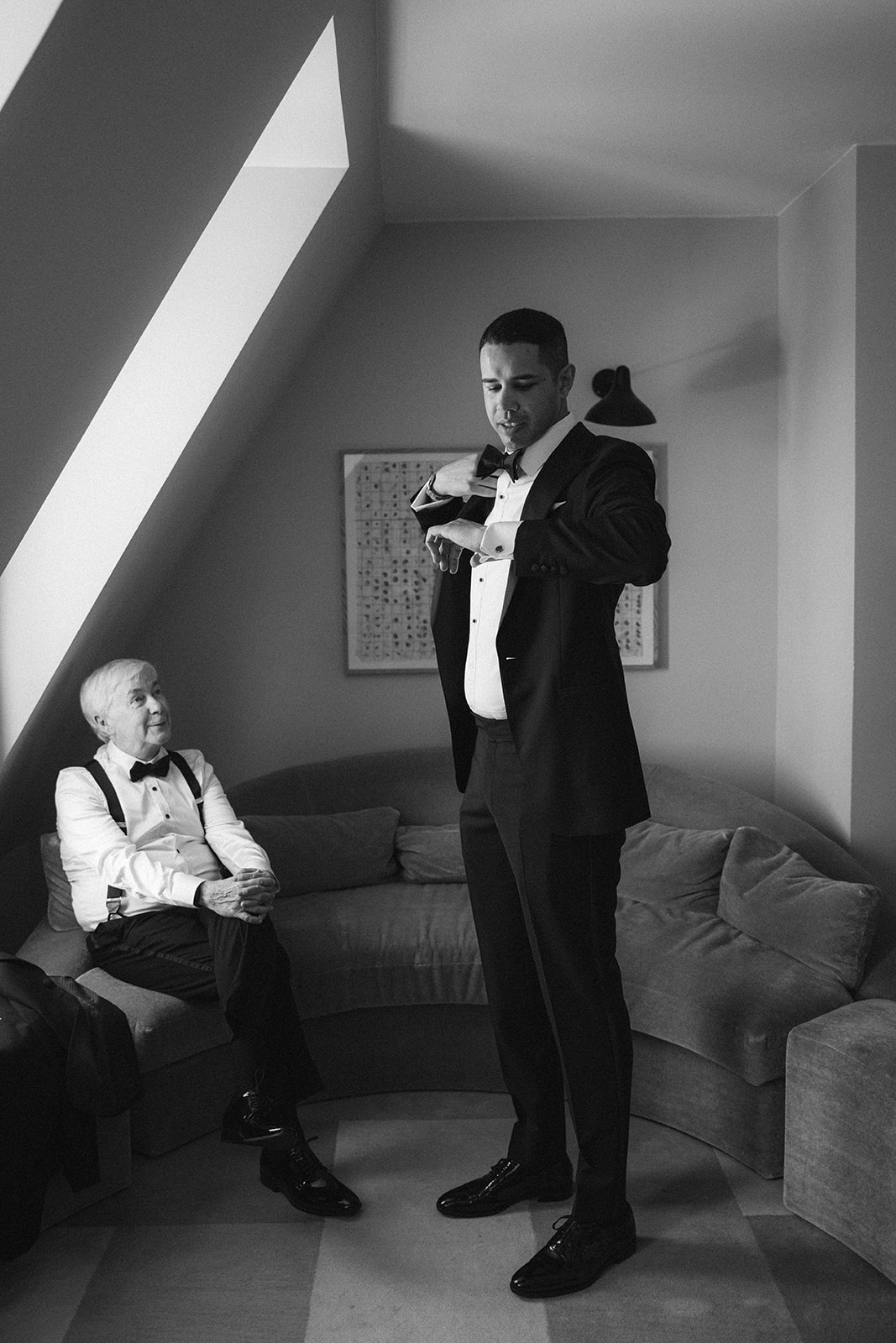 Black and white image of the groom adjusting his bow tie with his father seated nearby