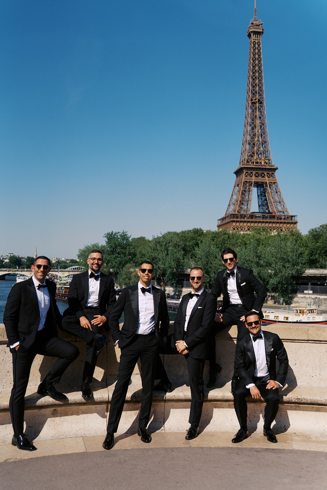 Six groomsmen and the groom in tuxedos pose on a Paris bridge with the Eiffel Tower behind
