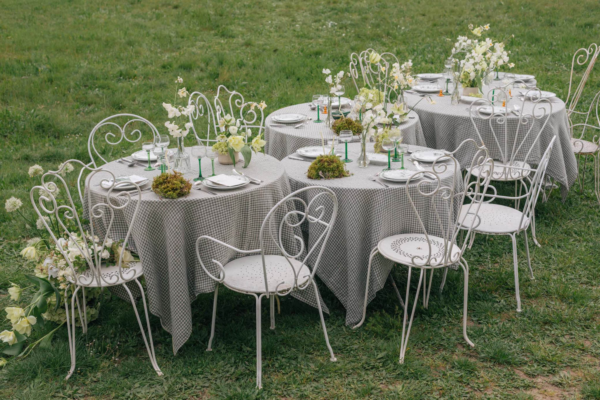 Round gingham-check tables with hellebore and allium bud vases and green-stemmed glasses on garden lawn