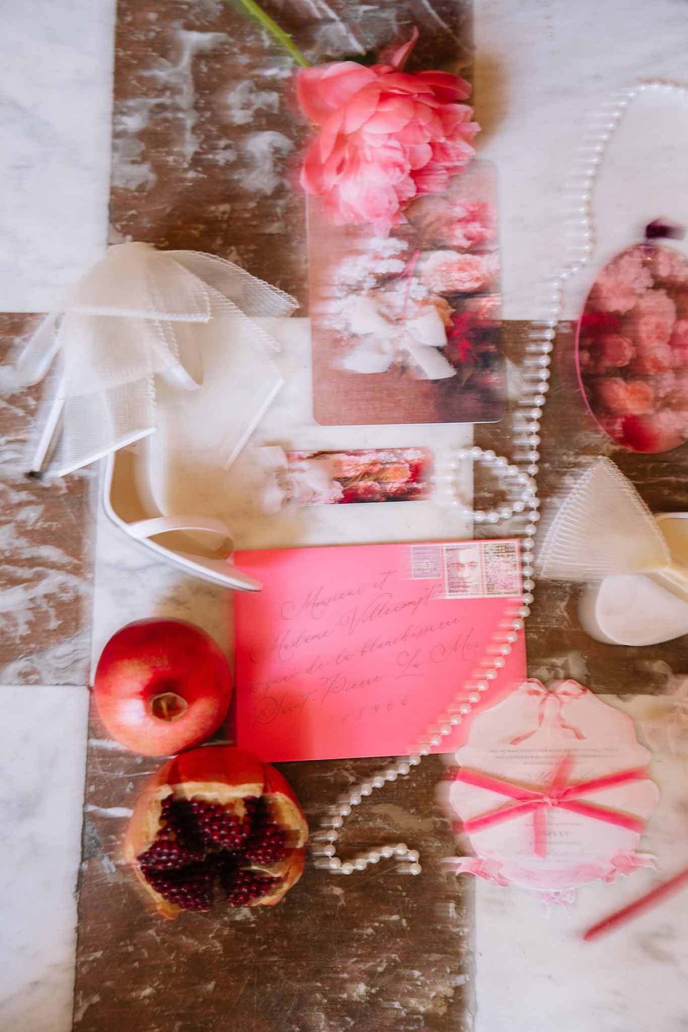 Flat lay of coral envelope, pink invitation suite, white satin heel, pearl strand, and coral peony on marble
