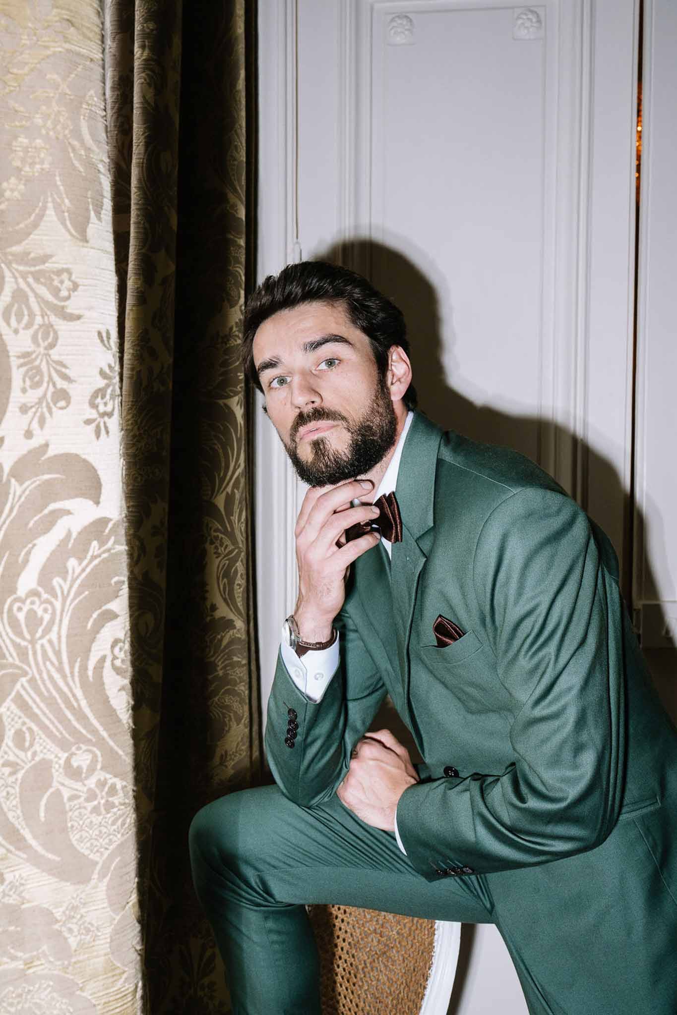 Groom in forest green double-breasted suit with burgundy bow tie posed beside gold damask curtain in panelled room