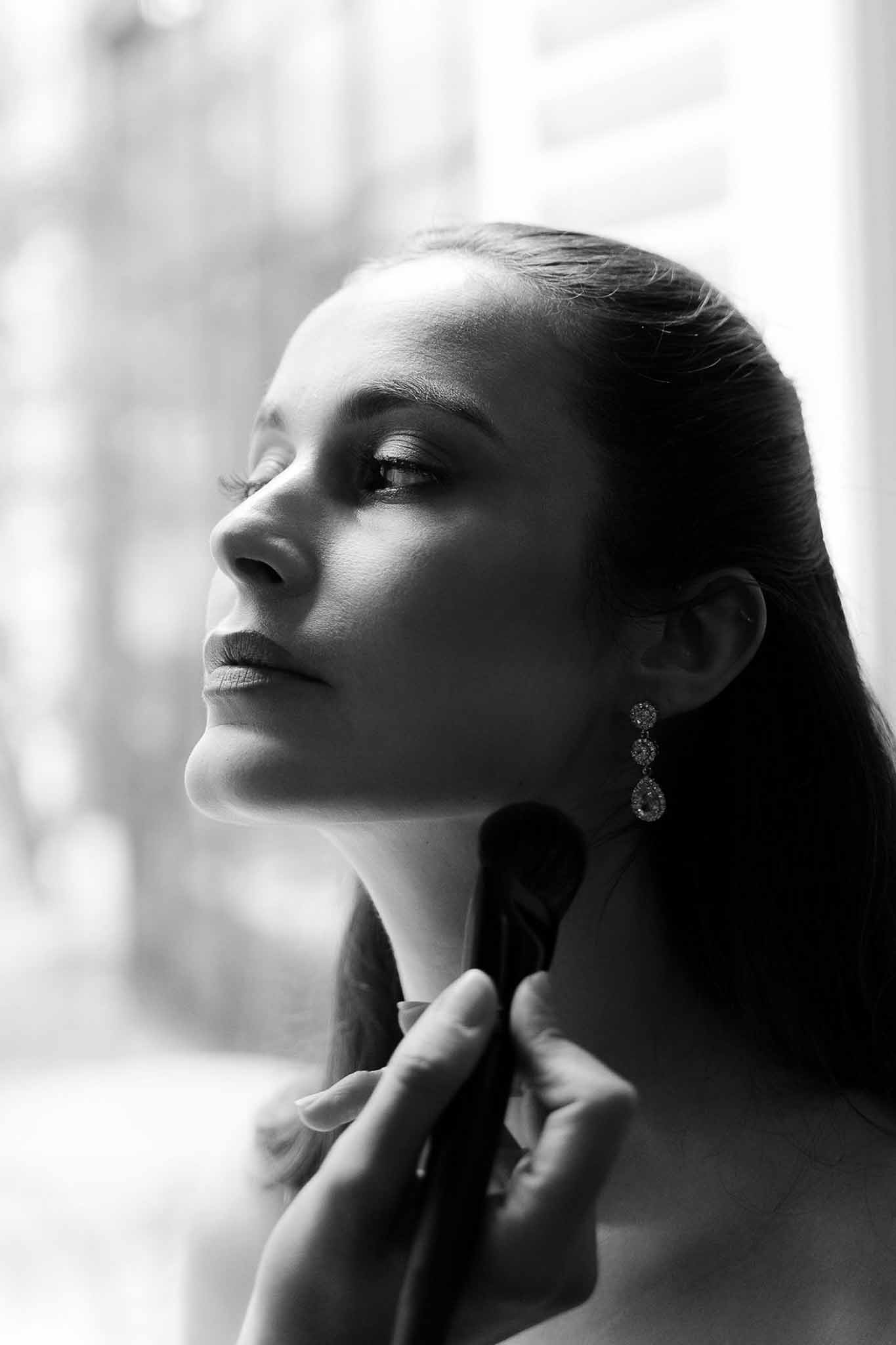 Black and white close-up of makeup artist applying powder to bride wearing crystal chandelier earring