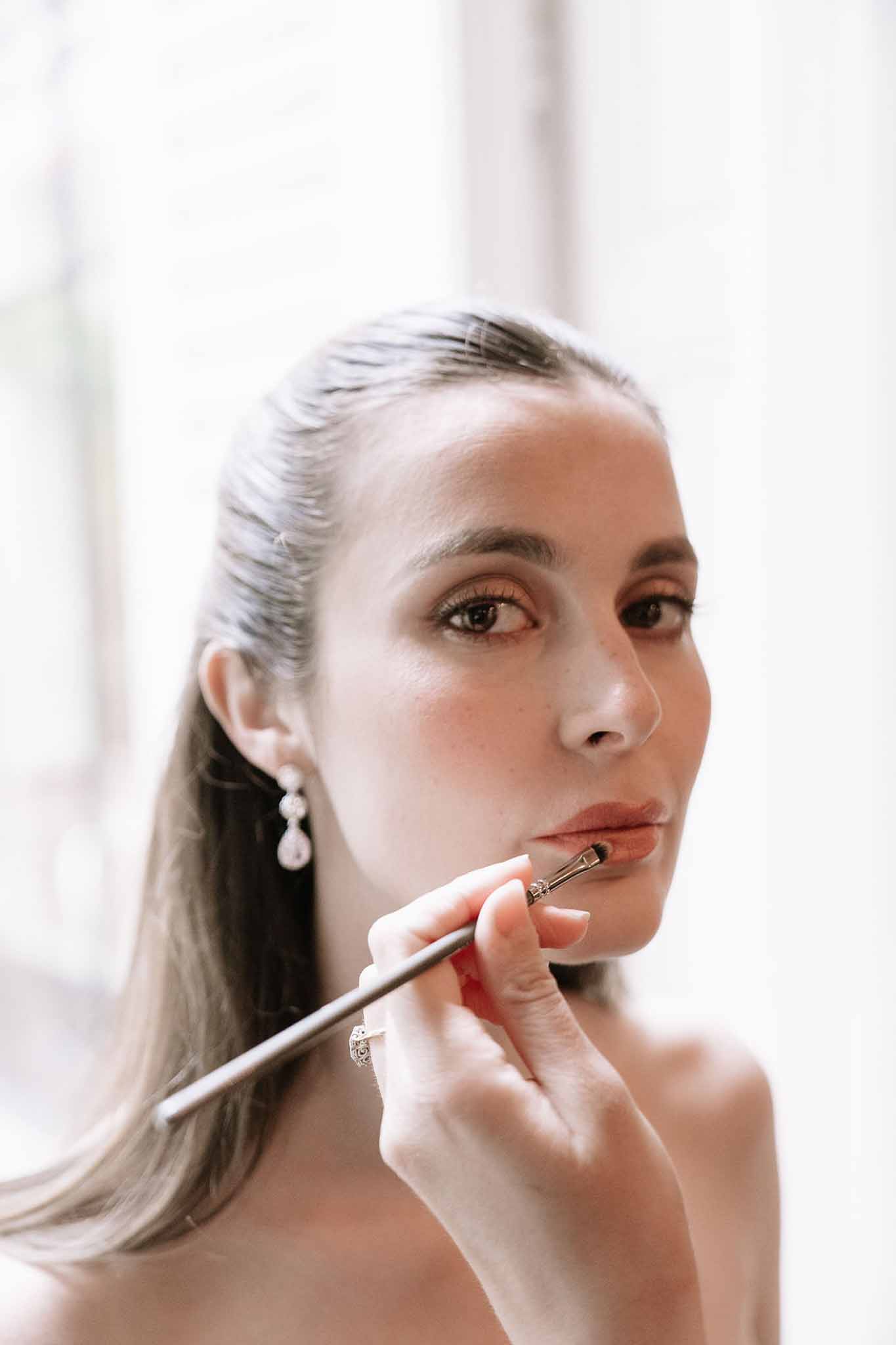 Close-up of bride having lip makeup applied with brush, sleek ponytail and crystal drop earrings