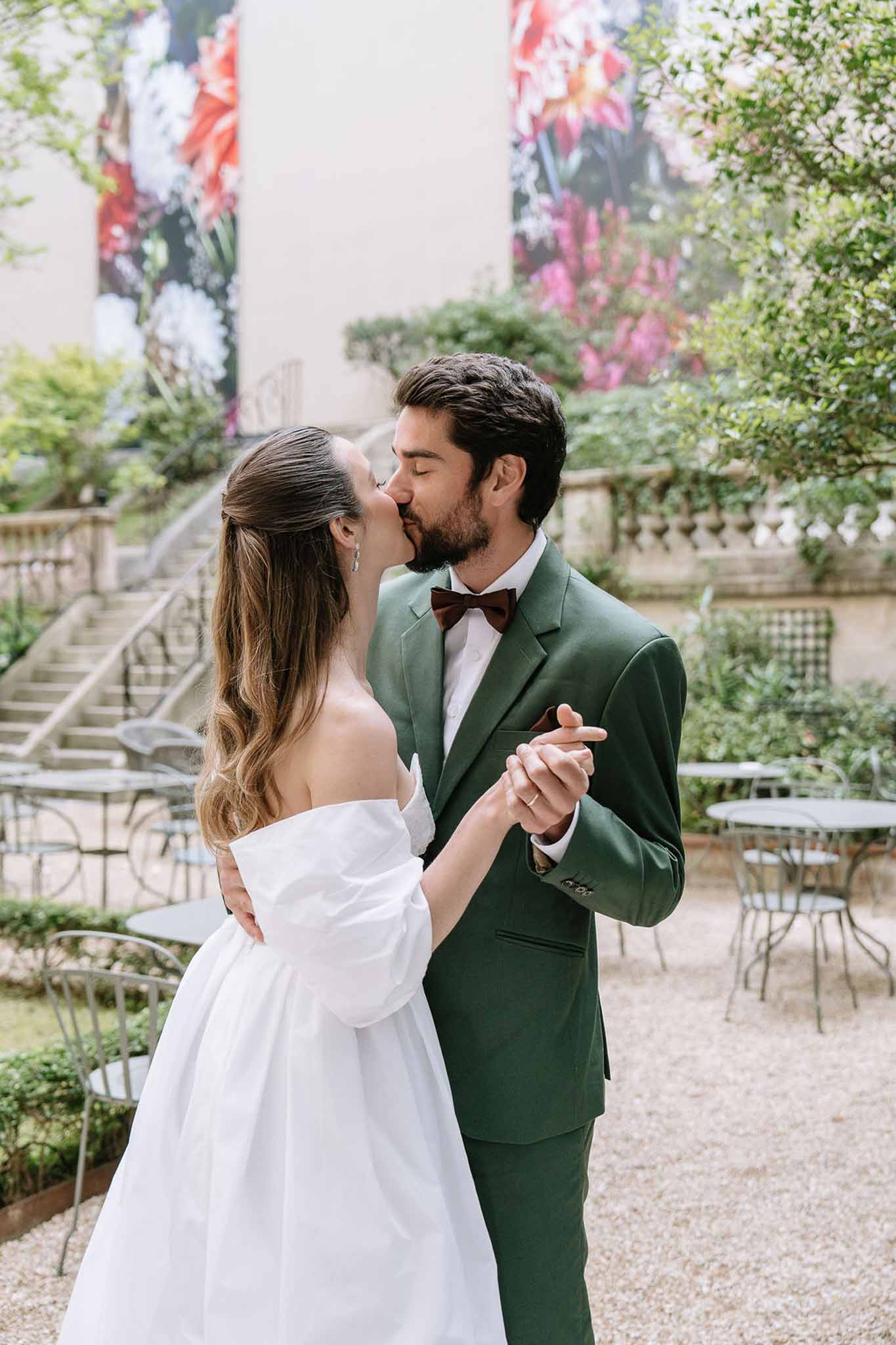 Couple kissing in courtyard with groom in forest green suit and floral print backdrop panels