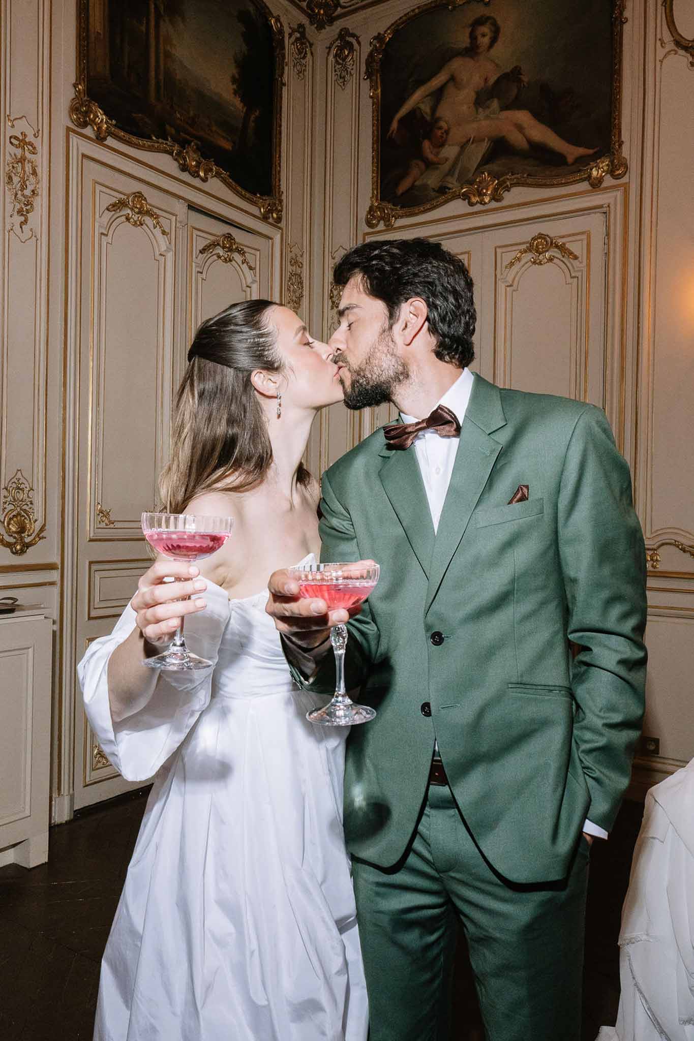Bride and groom kissing while holding pink cocktails in gilded French chateau reception room