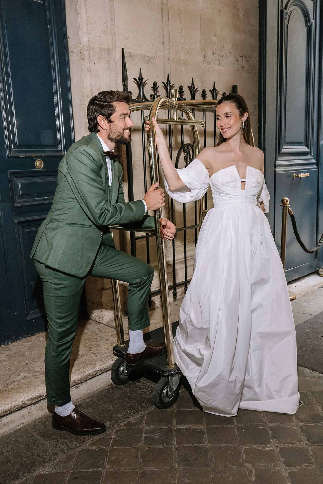Couple posing with brass luggage trolley outside Parisian hotel bride in ball gown groom in green suit