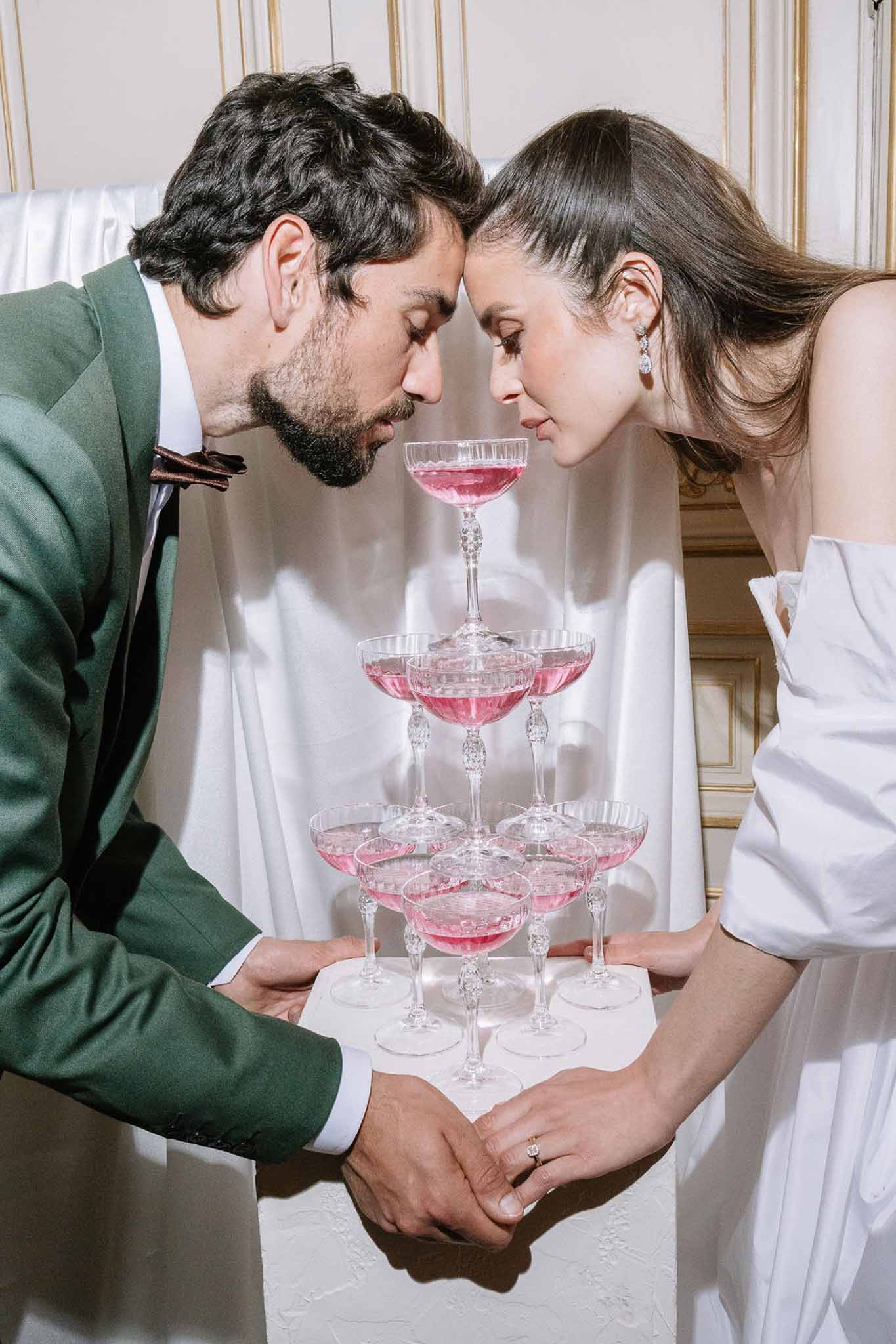 Couple leaning over four-tier pink champagne coupe tower with groom in green suit and burgundy bow tie