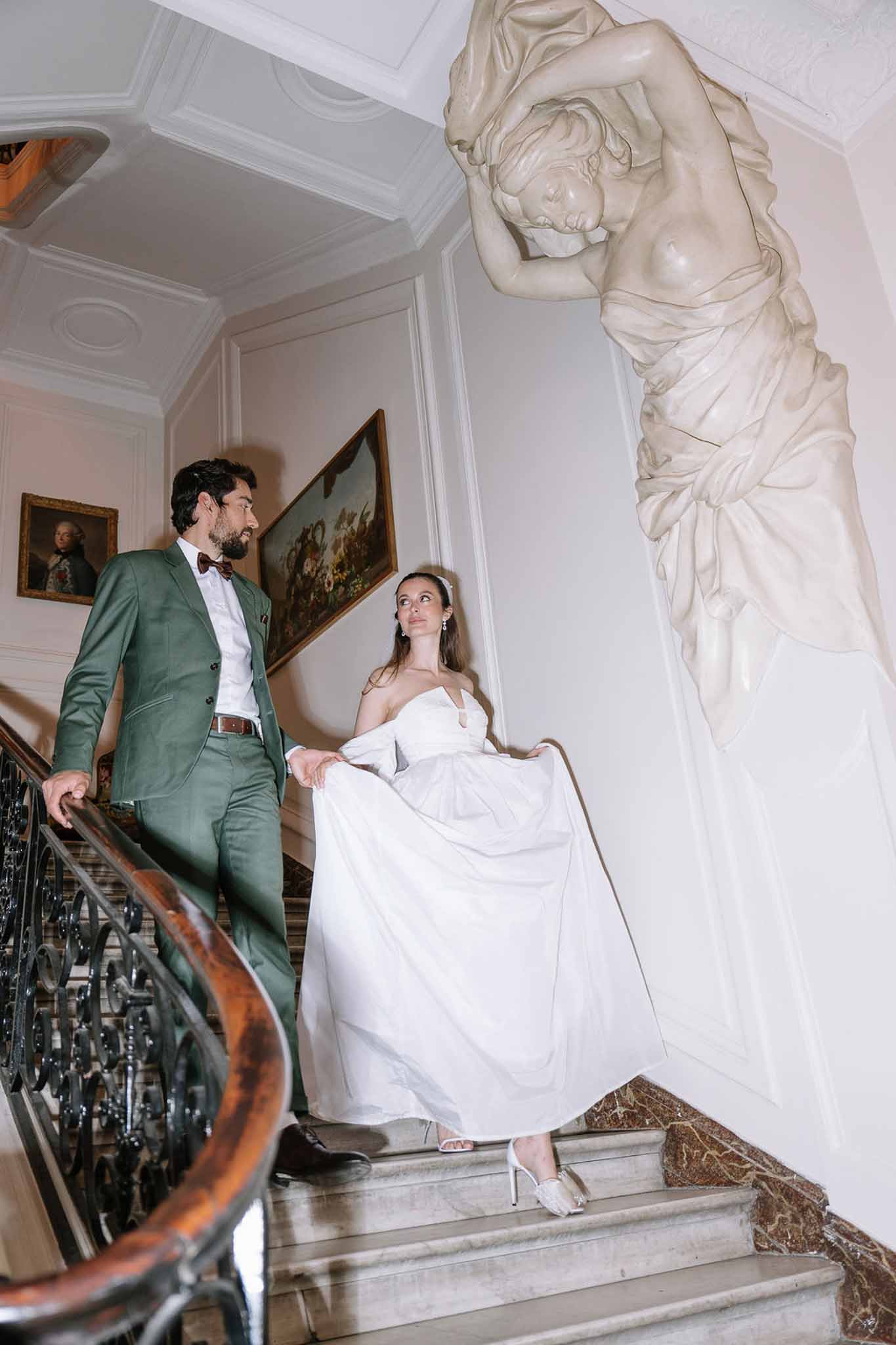 Bride descending marble staircase as groom in sage suit stands above beside classical sculpture and gilt paintings