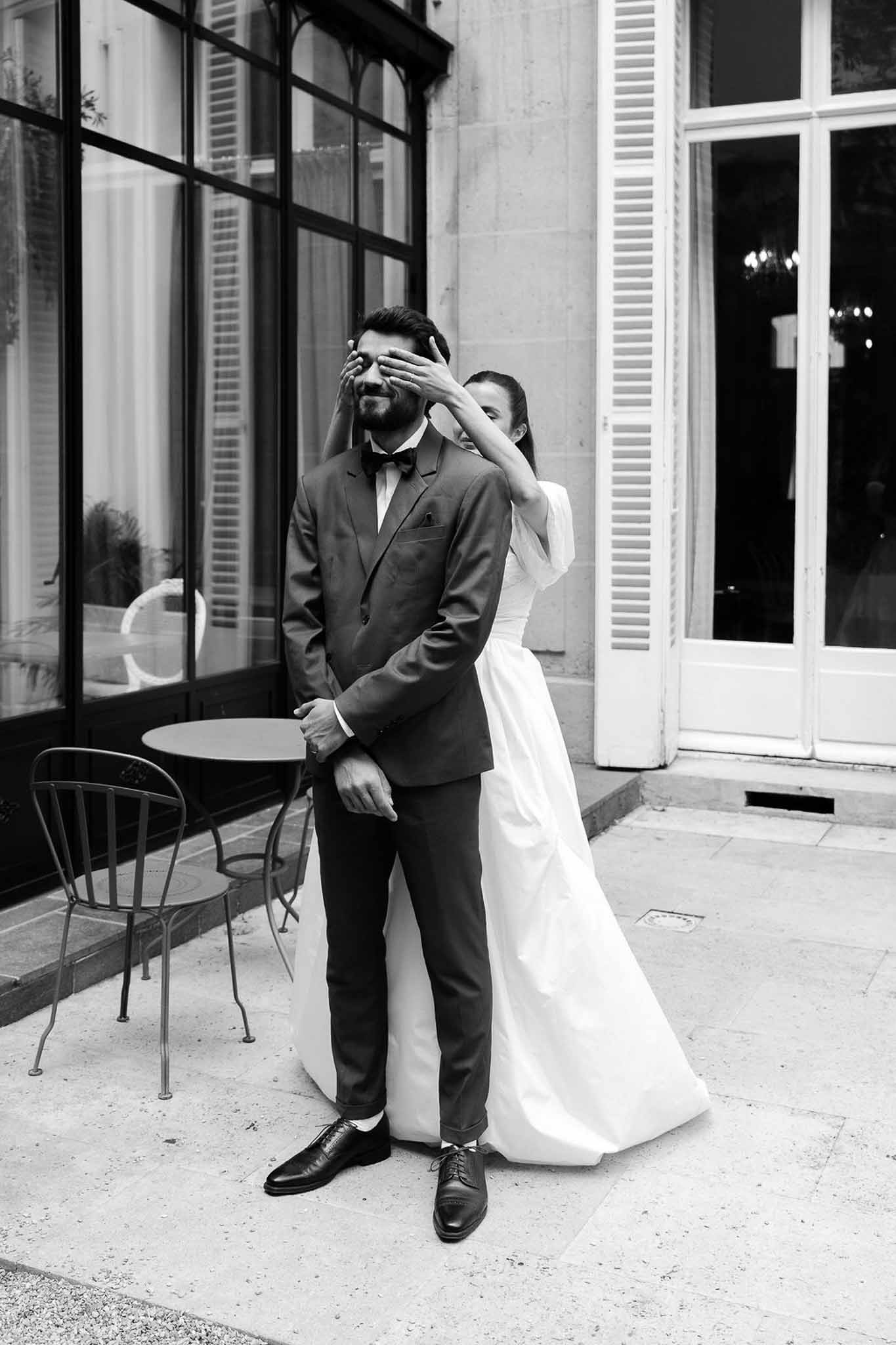 Bride in puff-sleeve gown covers groom's eyes from behind on stone terrace during first look in B&W
