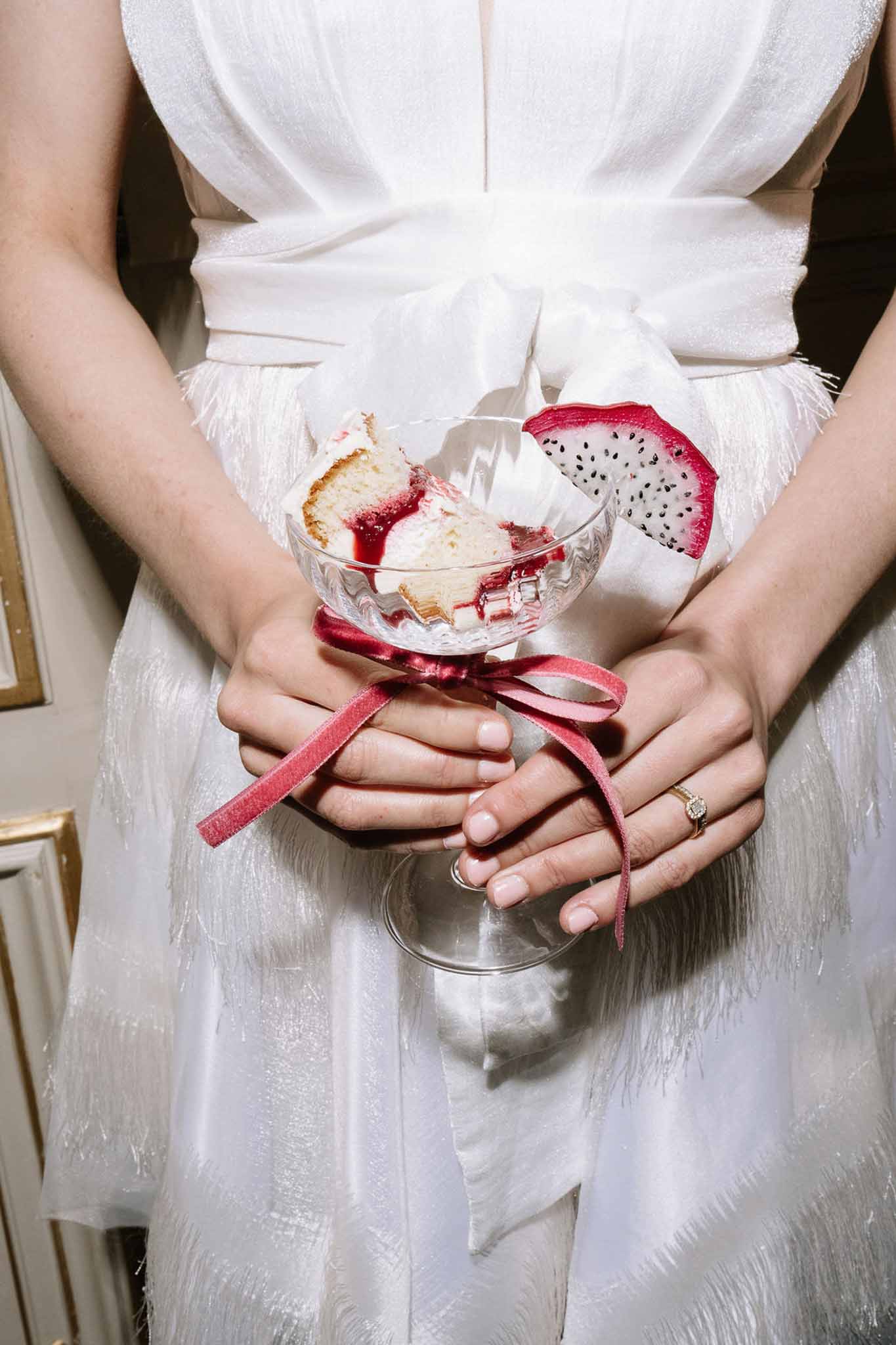 Bride holding coupe glass dessert with berry coulis and dragon fruit tied with crimson velvet ribbon