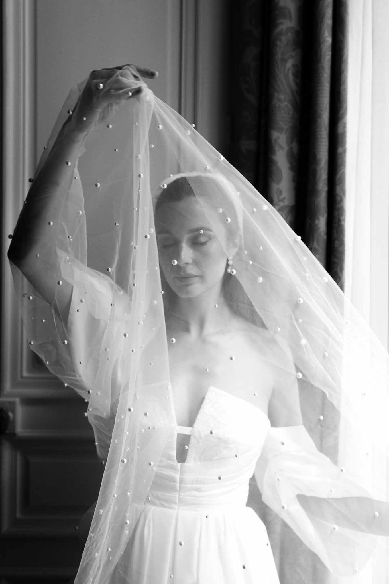 Bride holding pearl-embellished tulle veil above her head backlit by window in chateau room in B&W