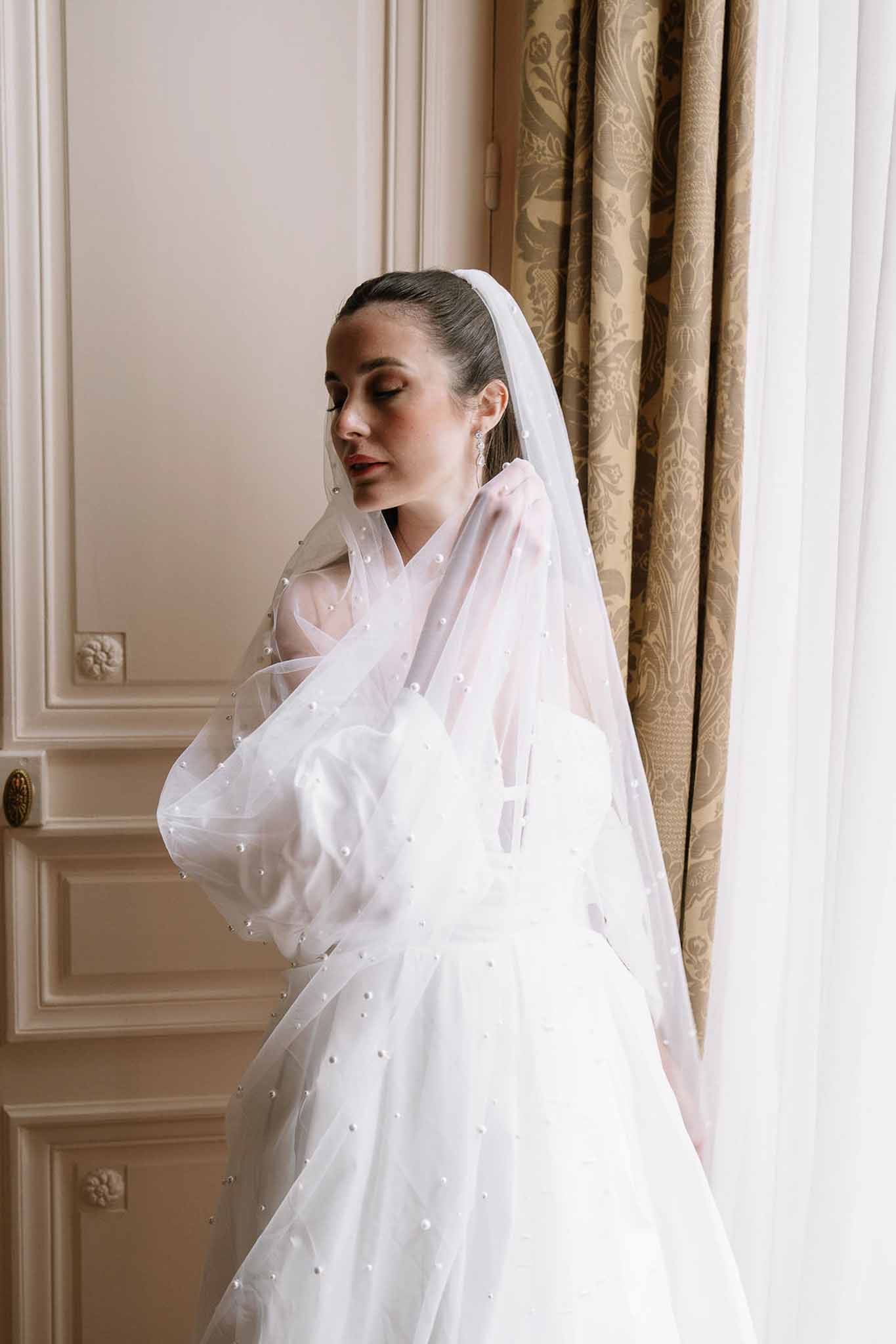 Bride in pearl-scattered veil and ballgown with crystal earrings by window in panelled hotel suite