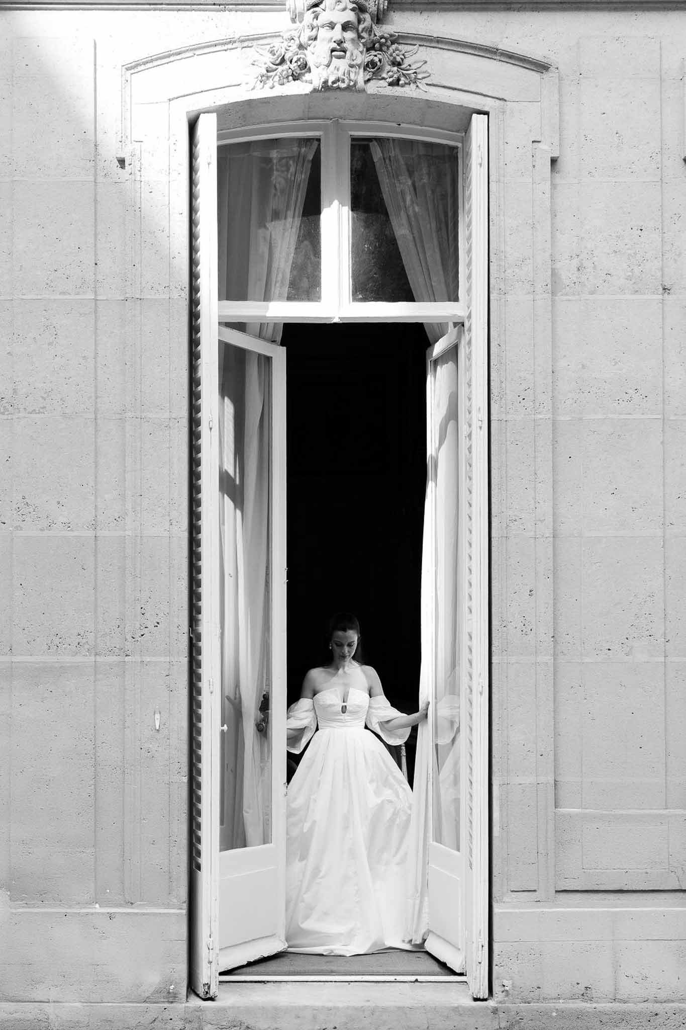 Bride in puff-sleeve ballgown standing in arched chateau doorway with carved stone mascaron above in B&W