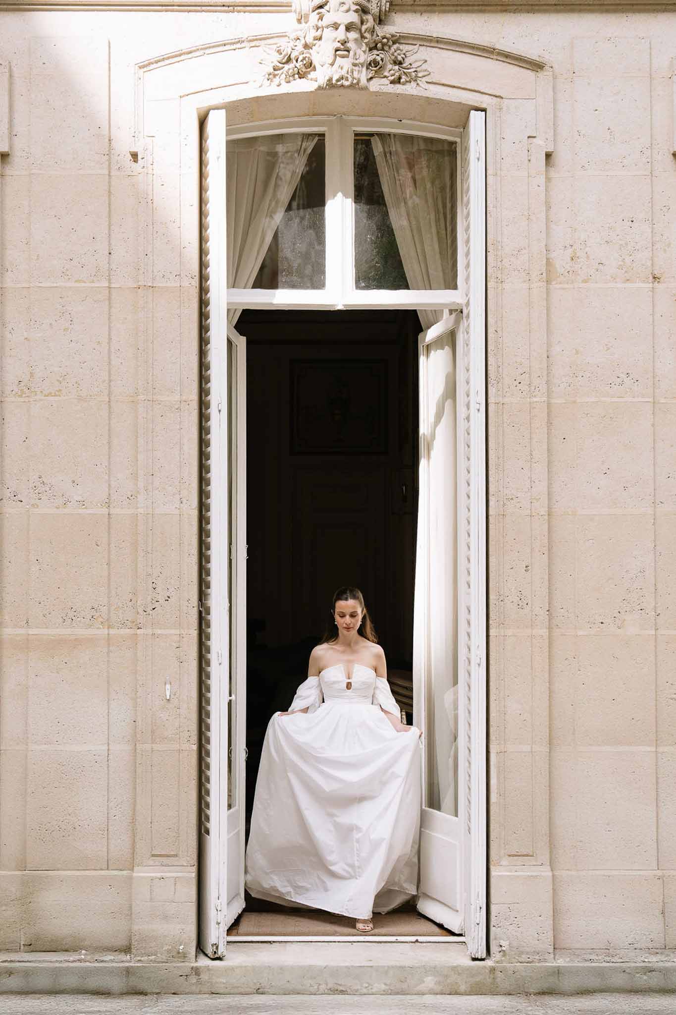 Bride in off-the-shoulder white ball gown standing in ornate stone chateau doorway with arched transom
