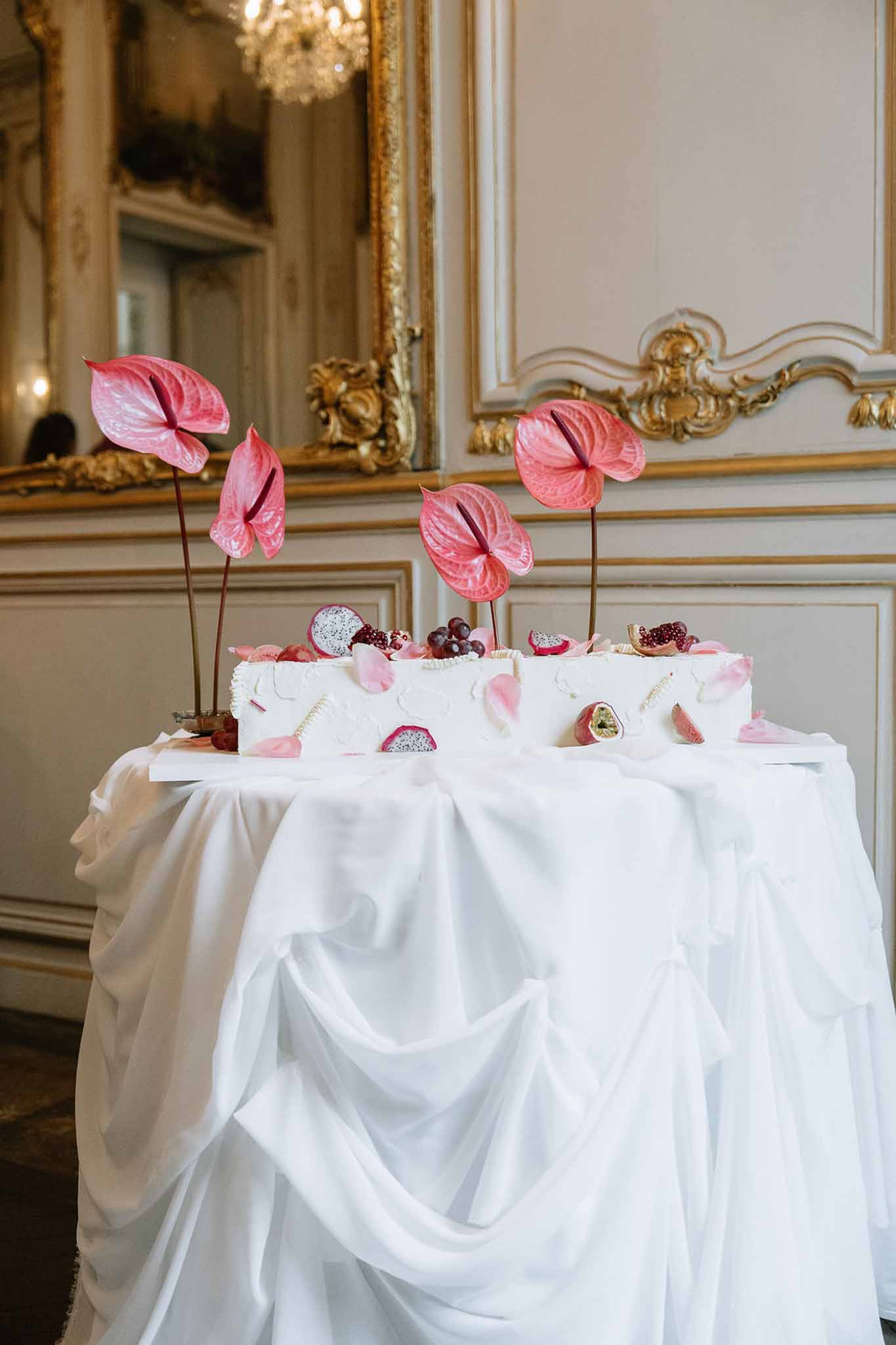 White wedding cake with tropical fruit and pink anthurium flowers on draped table in gilded French interior