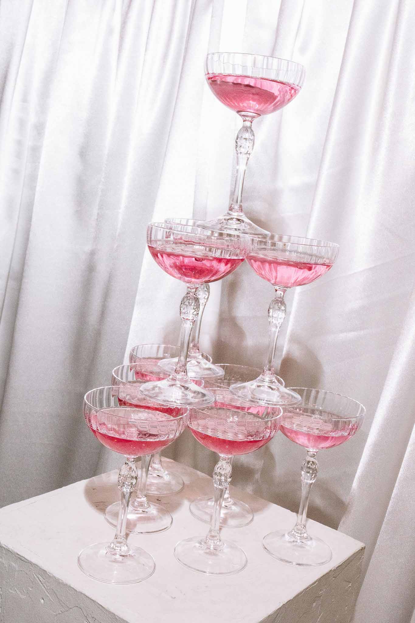Three-tier crystal coupe champagne tower filled with pink rose on white pedestal with draped backdrop