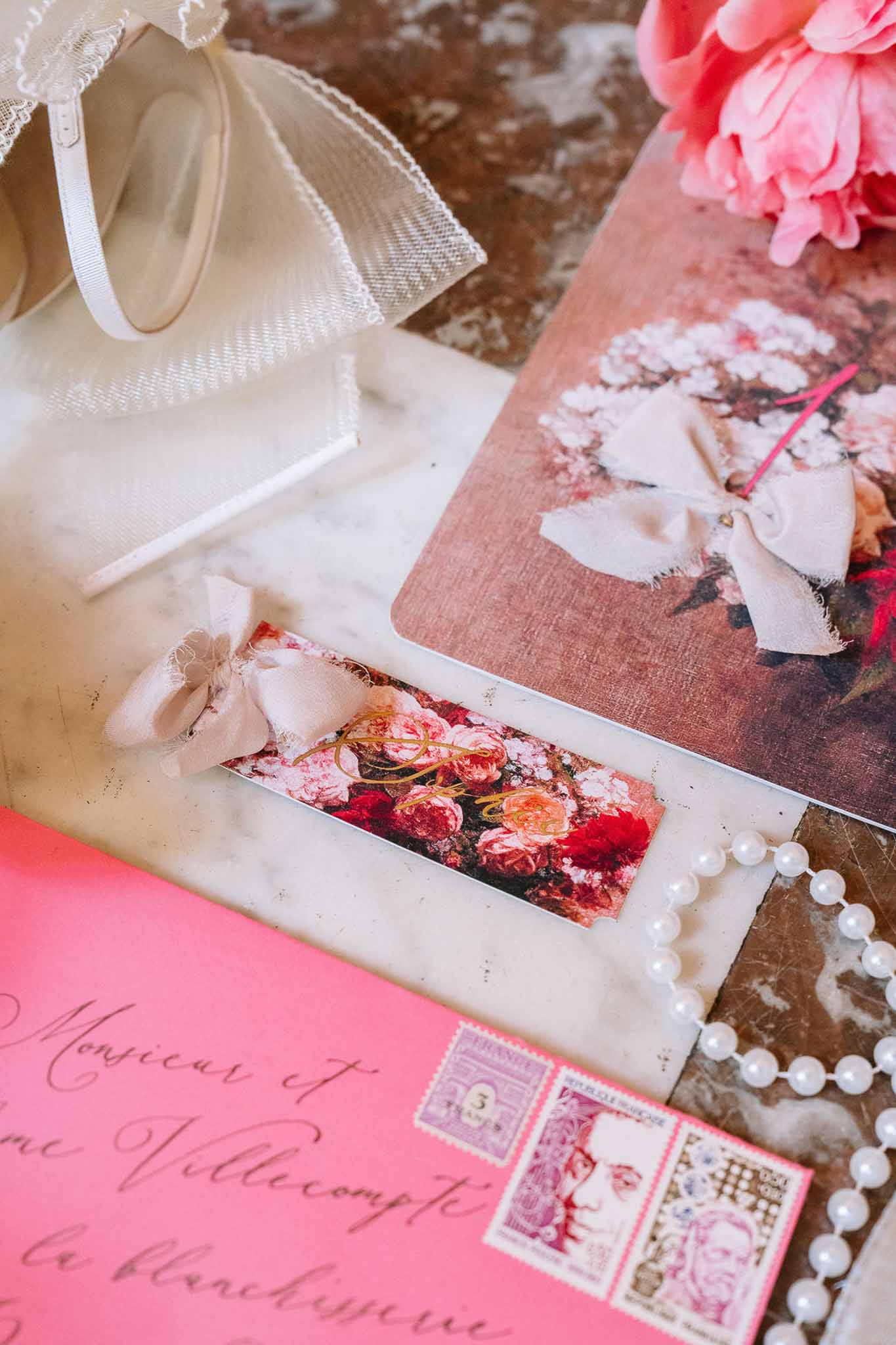 Coral pink envelope with vintage stamps, floral place cards, pearl necklace, and peony on marble