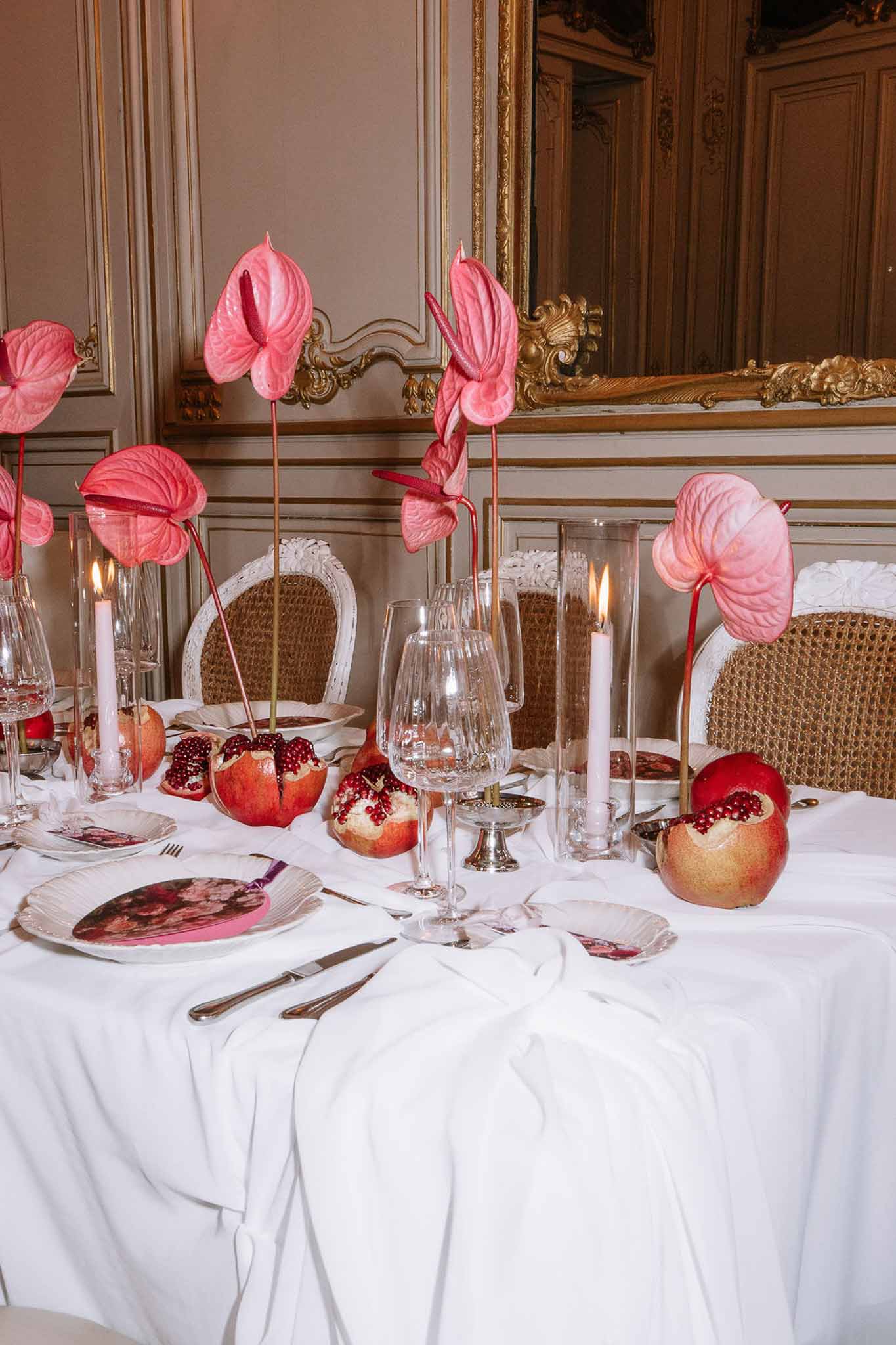 Hot pink anthurium stems with halved pomegranates and floral-printed chargers on white linen in gilt-paneled room