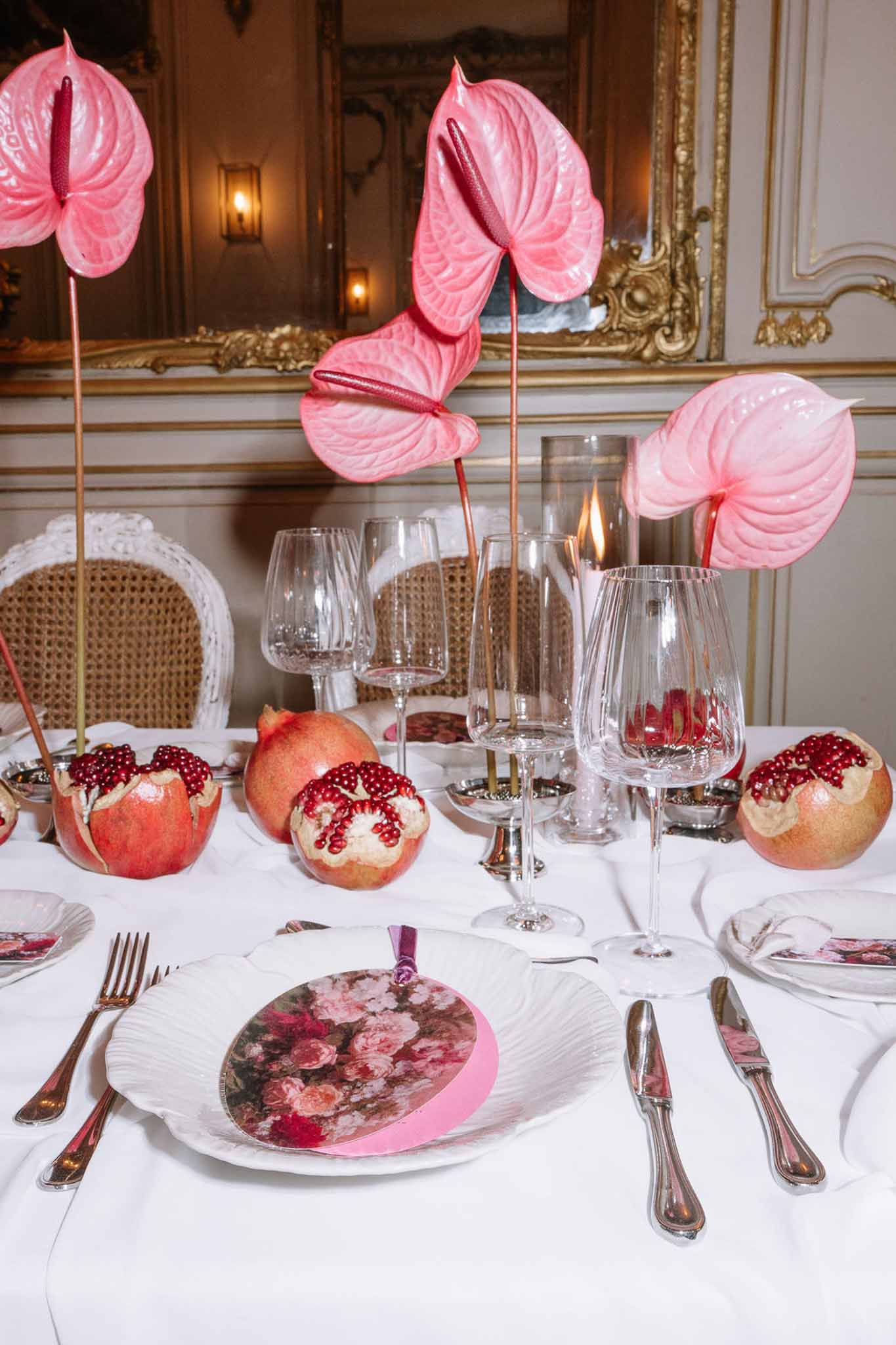 Reception table with pink anthurium centerpiece, pomegranates, floral menu cards, and crystal glasses in chateau interior