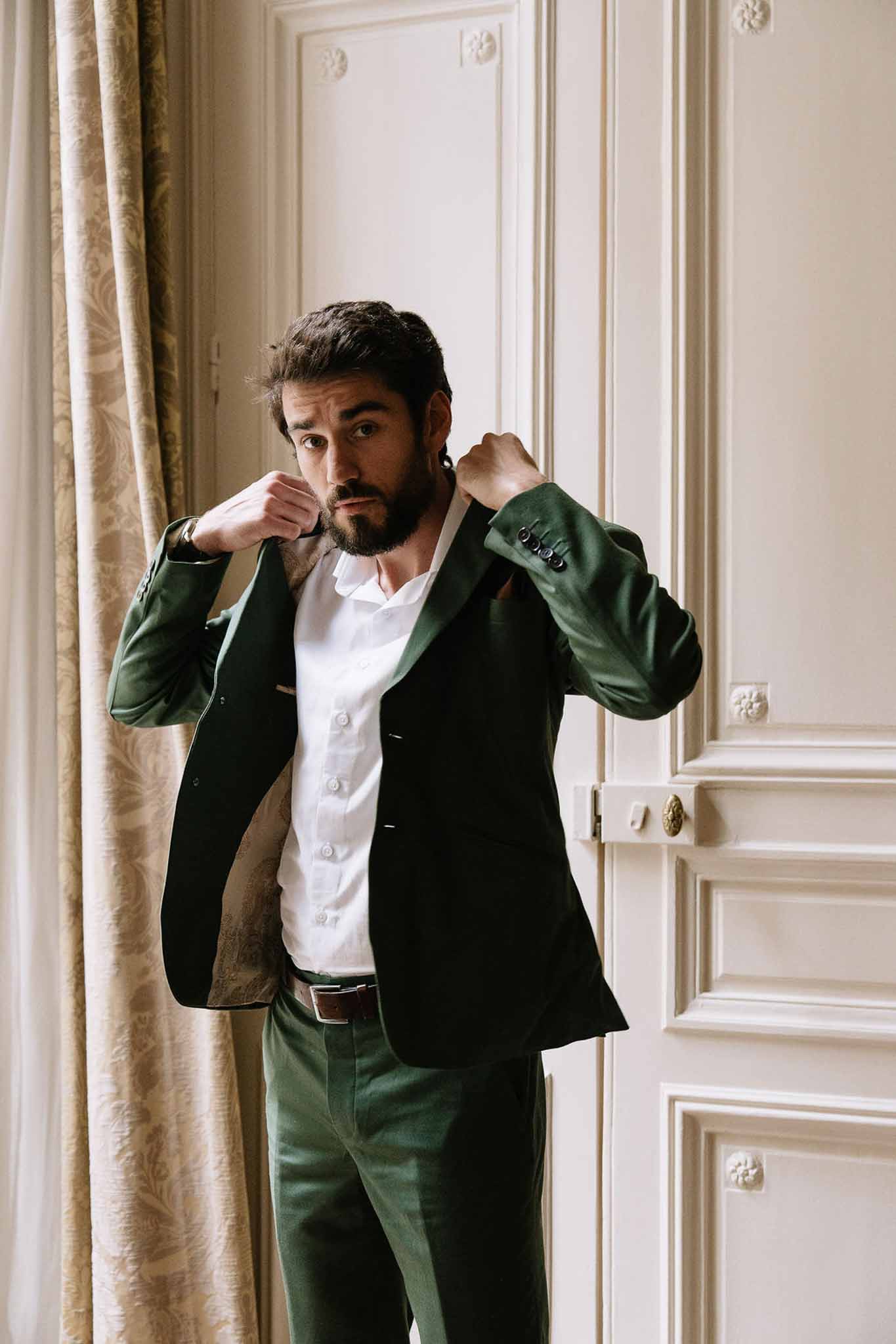 Groom adjusting forest green suit jacket before panelled door with damask curtain in chateau interior