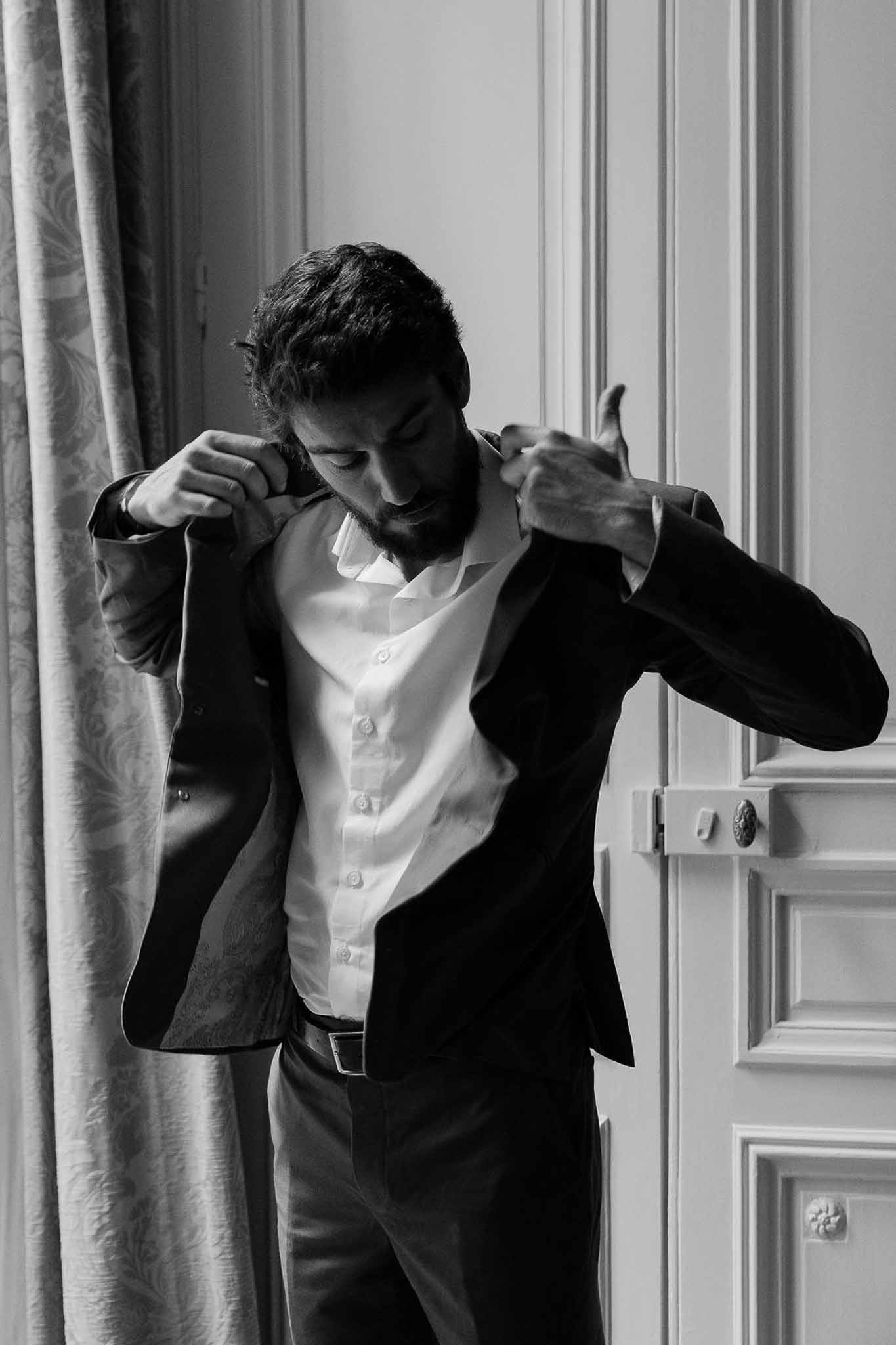 Black and white portrait of groom adjusting suit jacket in chateau room with panelled doors