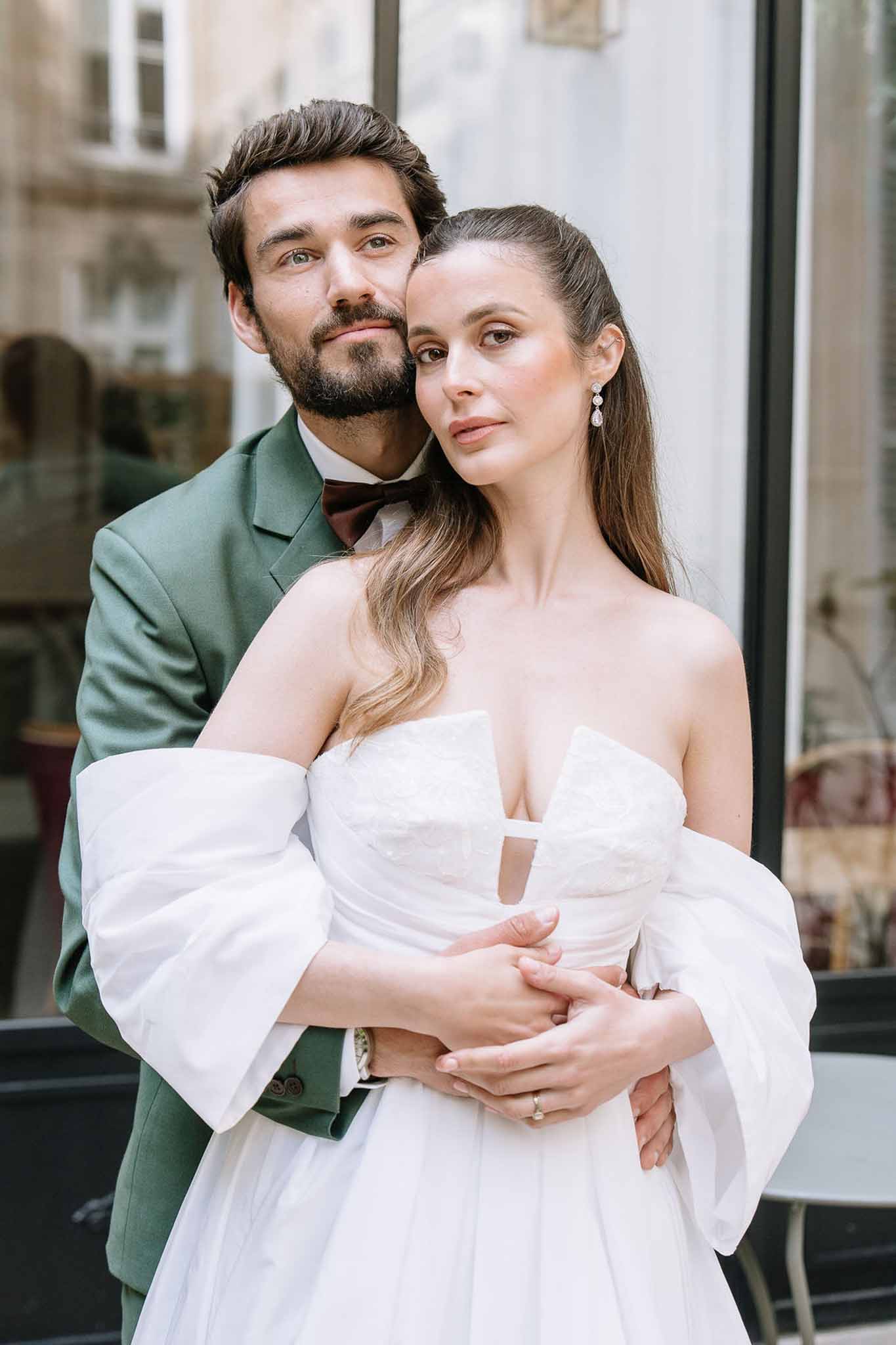 Groom in sage green suit embracing bride in off-shoulder white gown with puff sleeves in urban setting