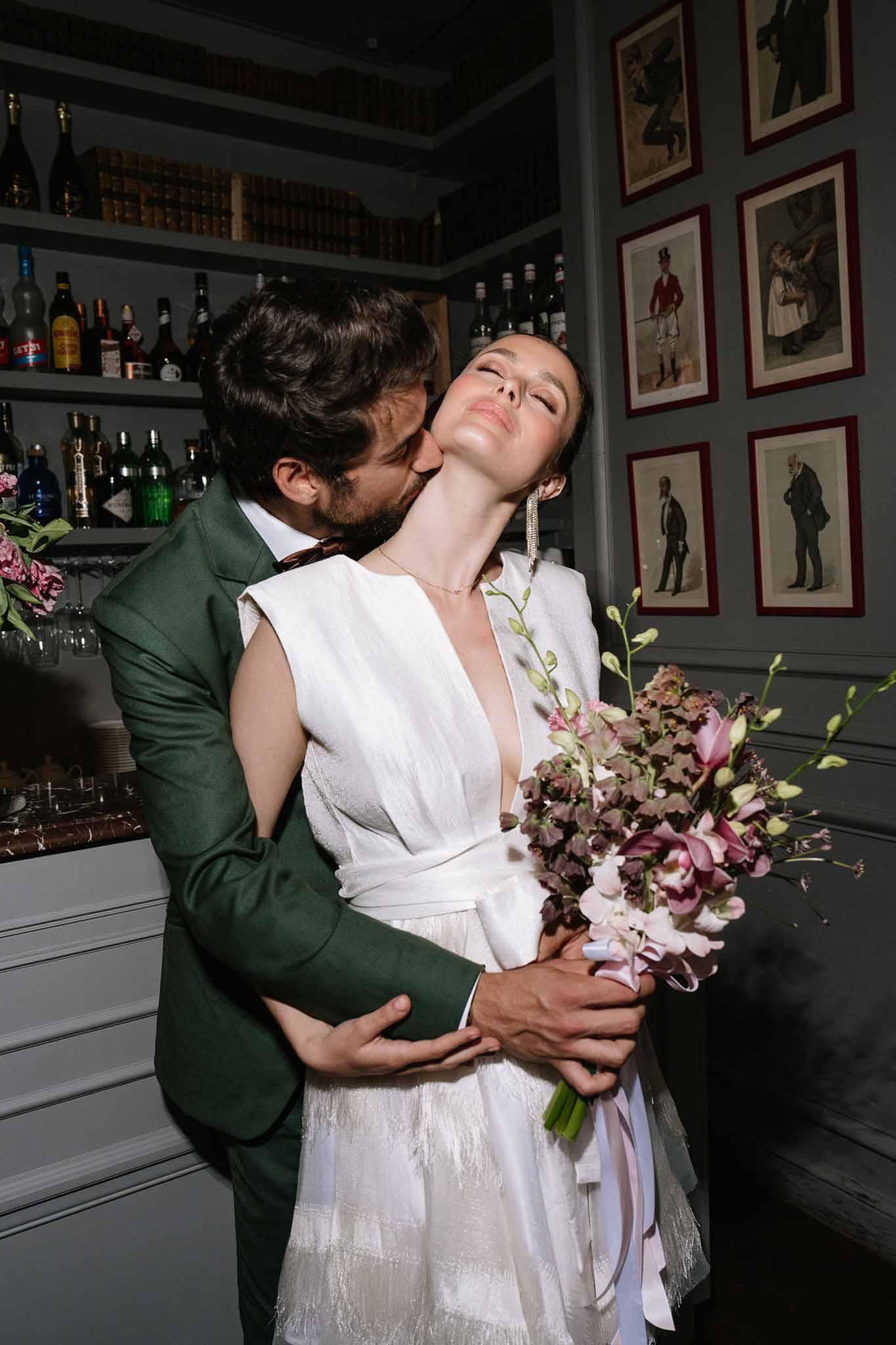 Groom in green suit kissing bride's neck as she holds dusky pink hellebore and orchid bouquet at bar