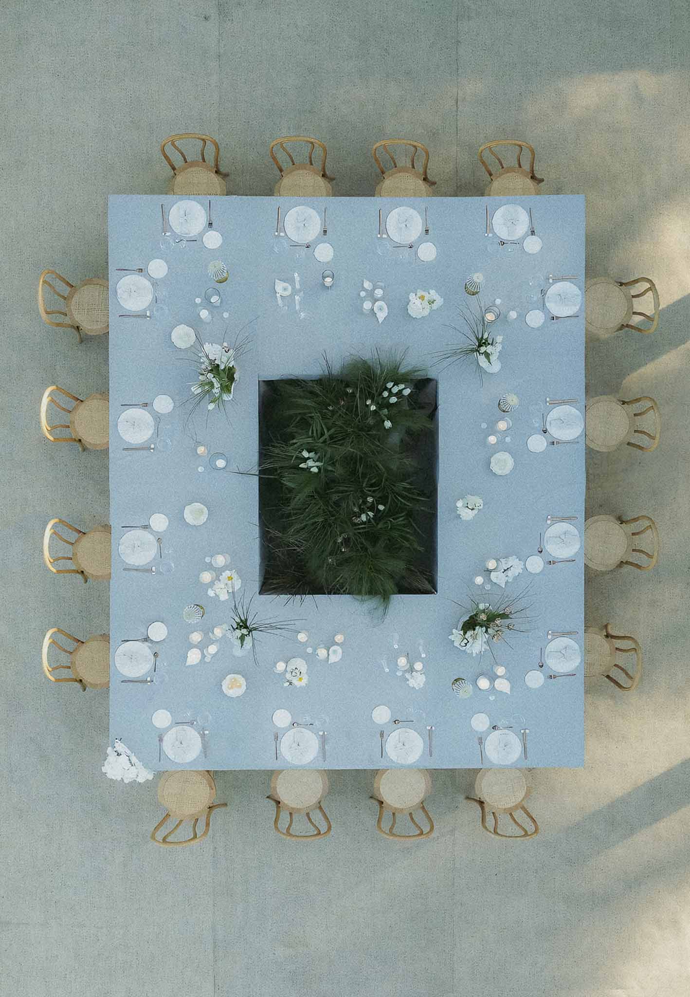 Aerial flat lay of square reception table with blue linen, feathery grasses, and gold rattan chairs