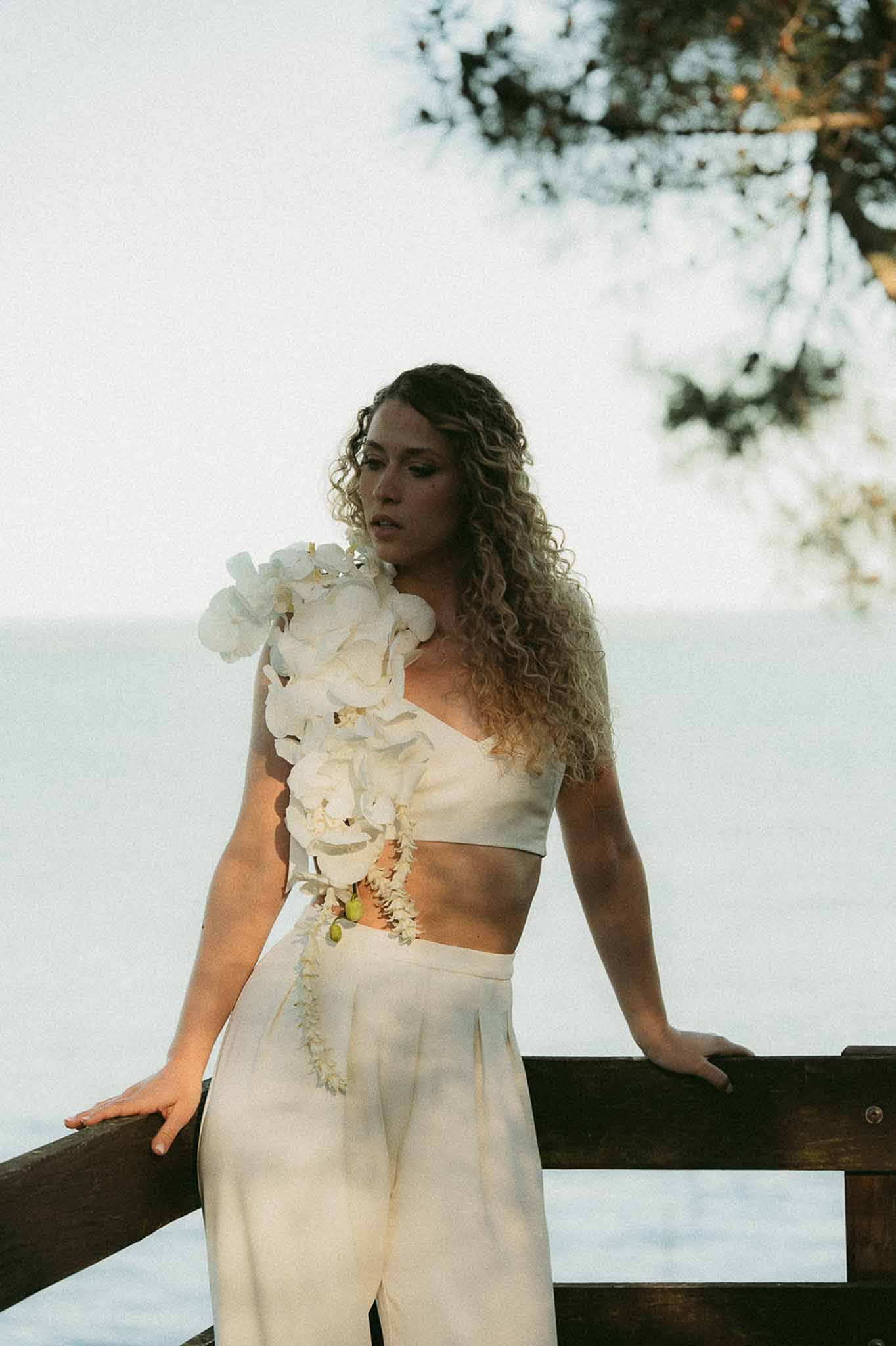 Bride in ivory crop top and wide-leg trousers with cascading orchid shoulder arrangement on waterfront deck